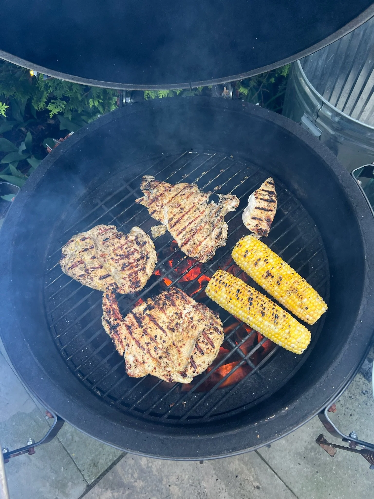 Grilled corn on the cob and several pieces of grilled meat on a barbecue grill with open lid.