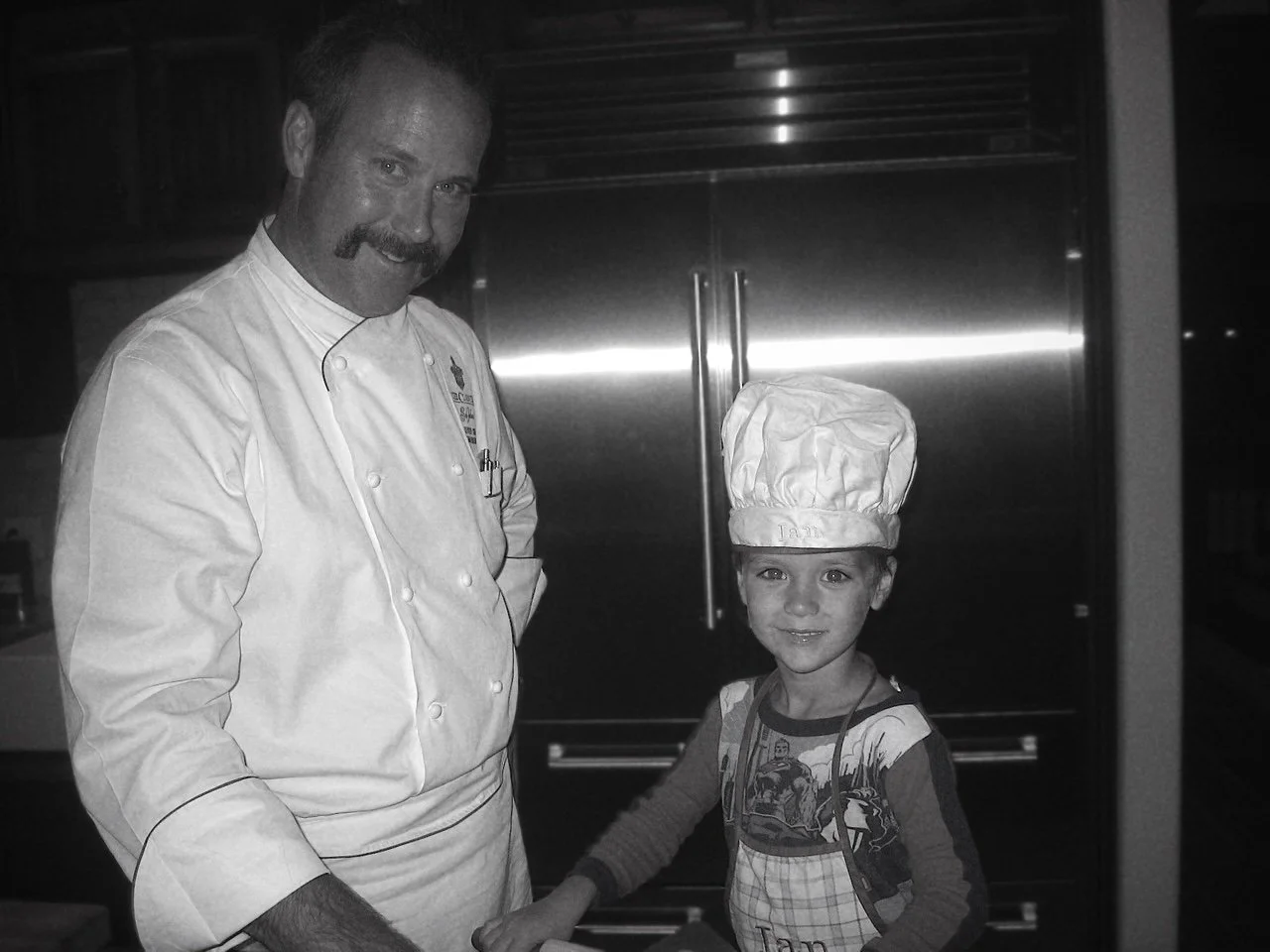 A chef and a young child wearing a chef's hat shake hands in a kitchen.