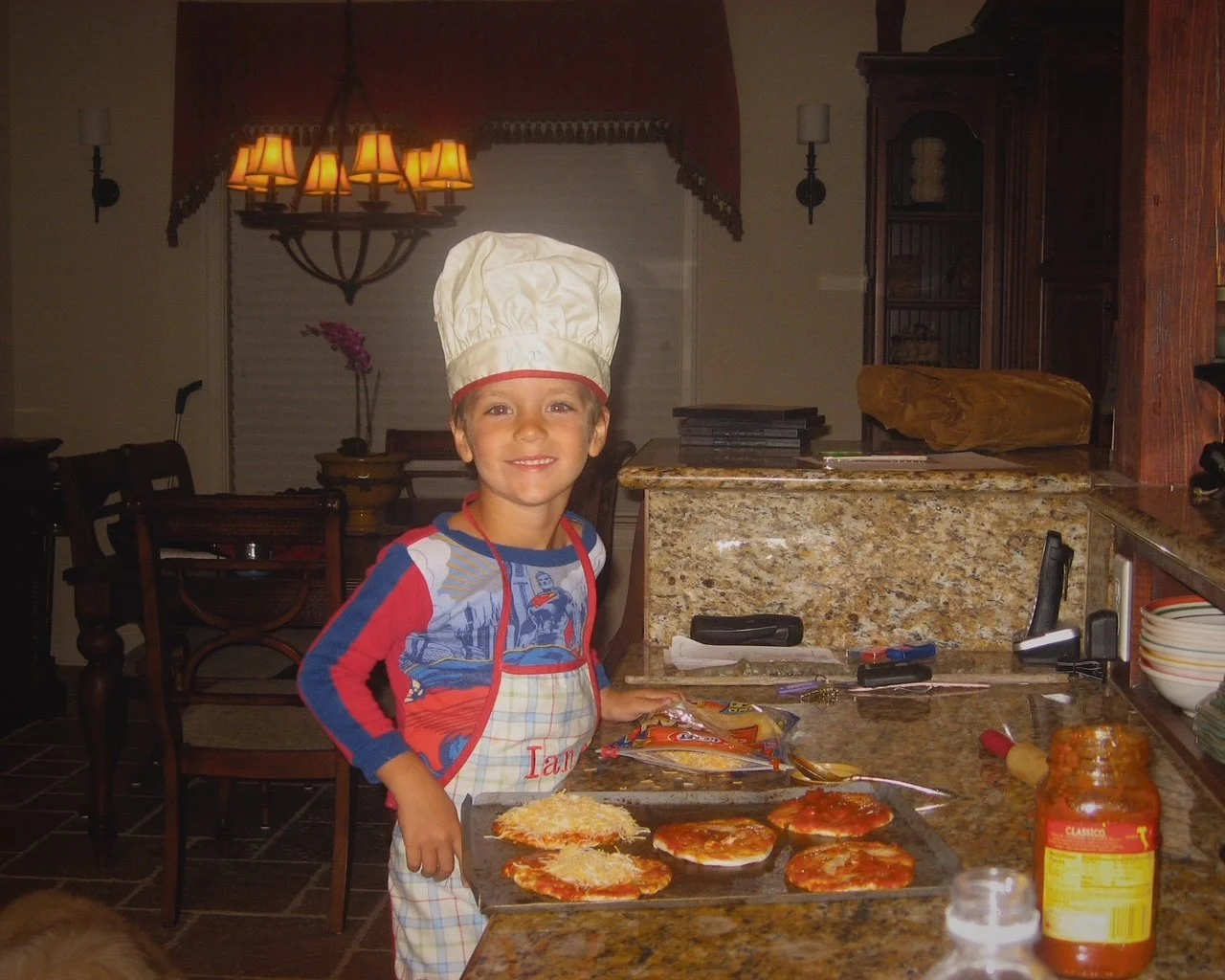 Child wearing a chef's hat and apron standing by a kitchen counter with pizza baking on a tray.