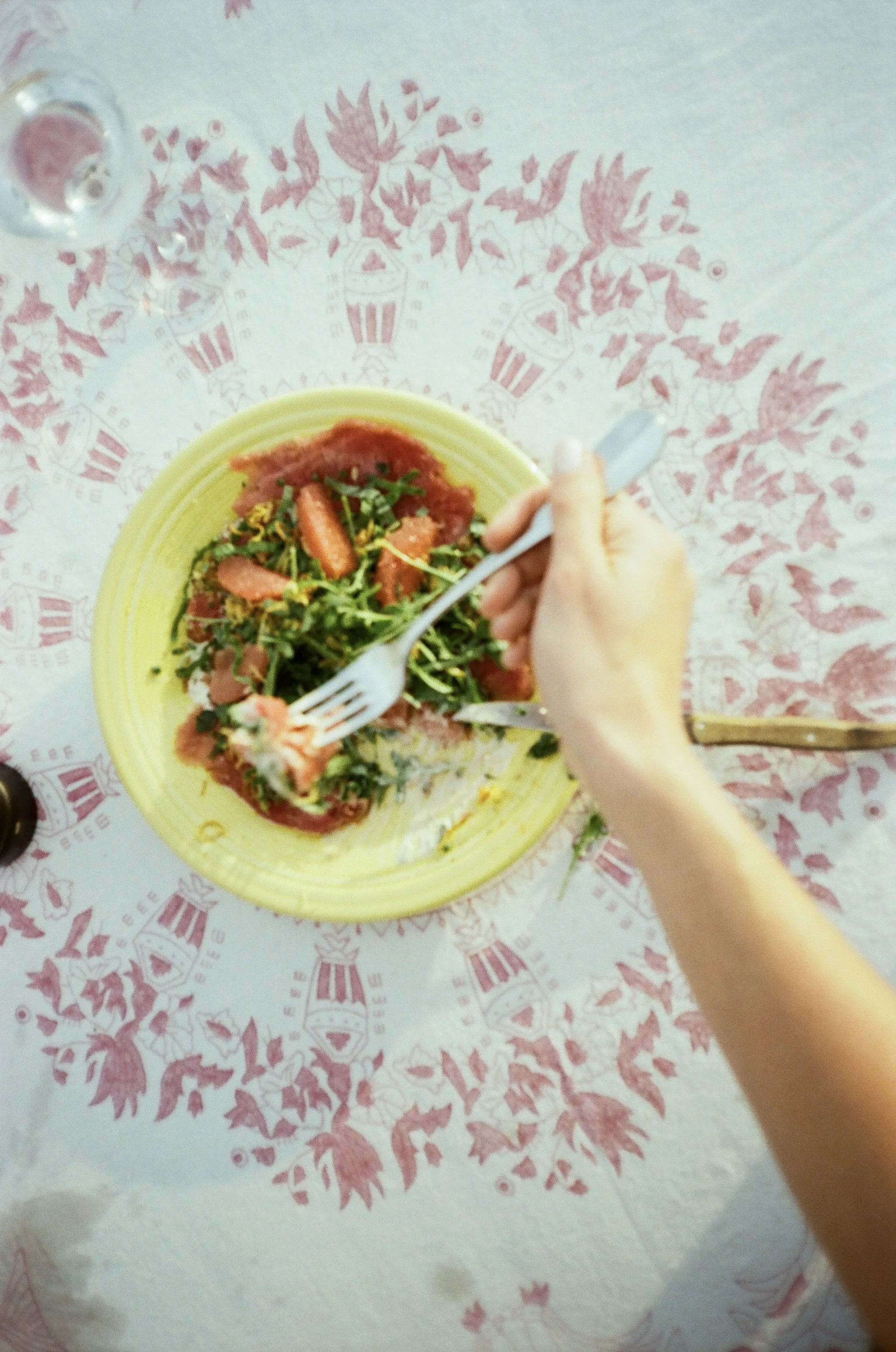 Person eating a salad with a fork and knife on a yellow plate, on a decorative tablecloth.
