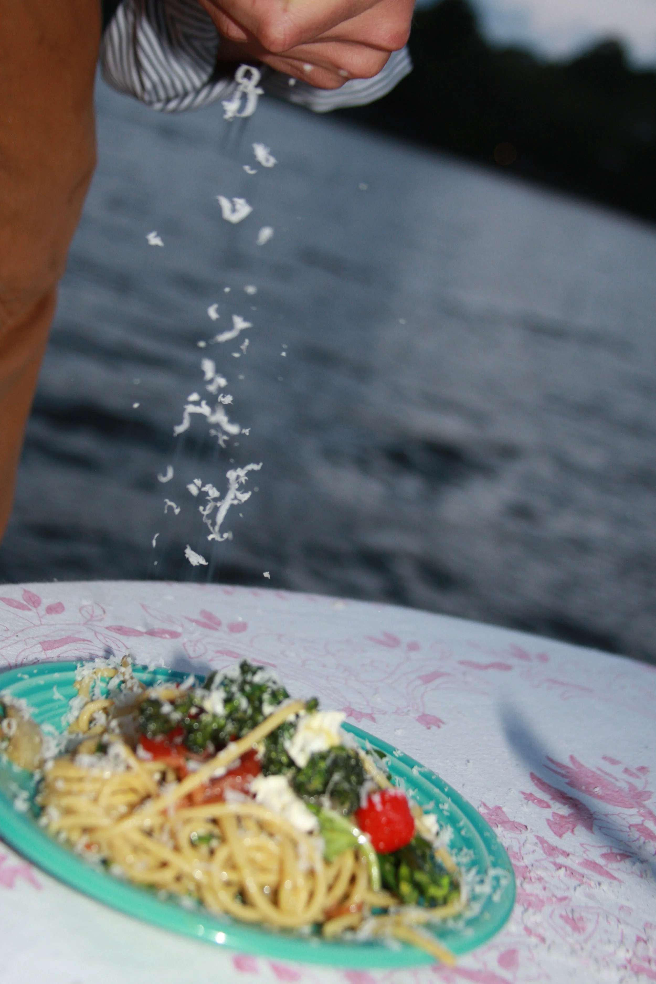 A person sprinkling salt onto a plate of spaghetti with vegetables, with a body of water in the background.