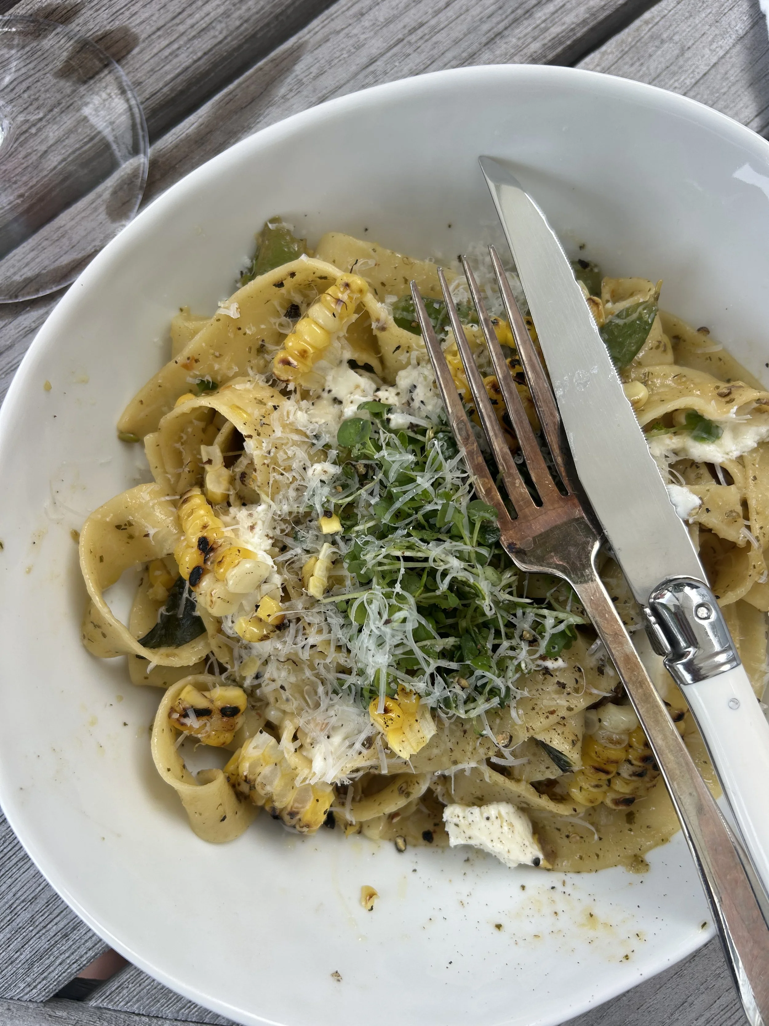 A white bowl of pasta with grilled corn, microgreens, and grated cheese on top, placed on a wooden table with a fork and knife resting on the edge.