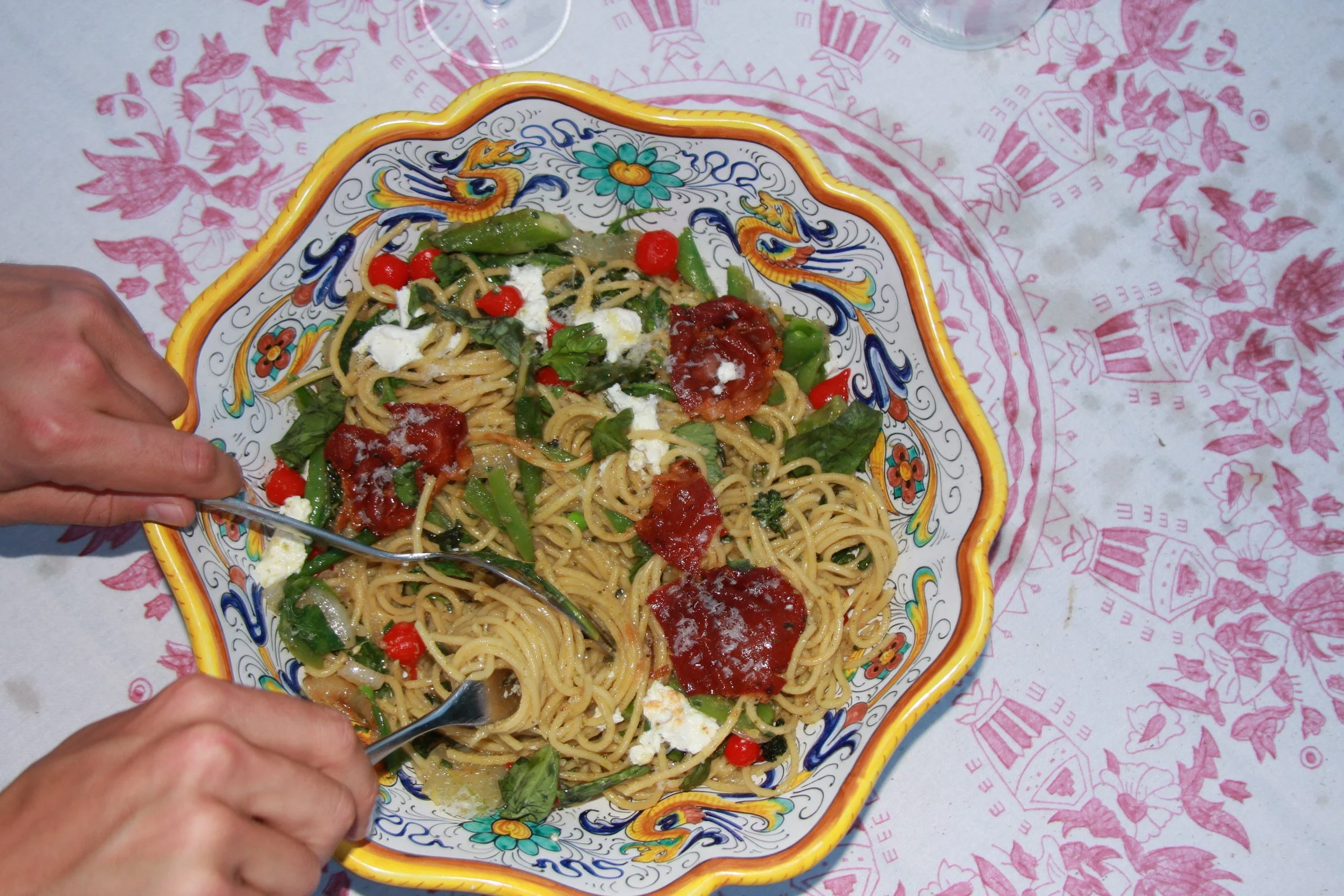 A colorful ceramic bowl of spaghetti with red sauce, cherry tomatoes, green peppers, greens, and white cheese. Two hands are holding a fork and spoon, eating the pasta at a table with a pink and white patterned tablecloth.