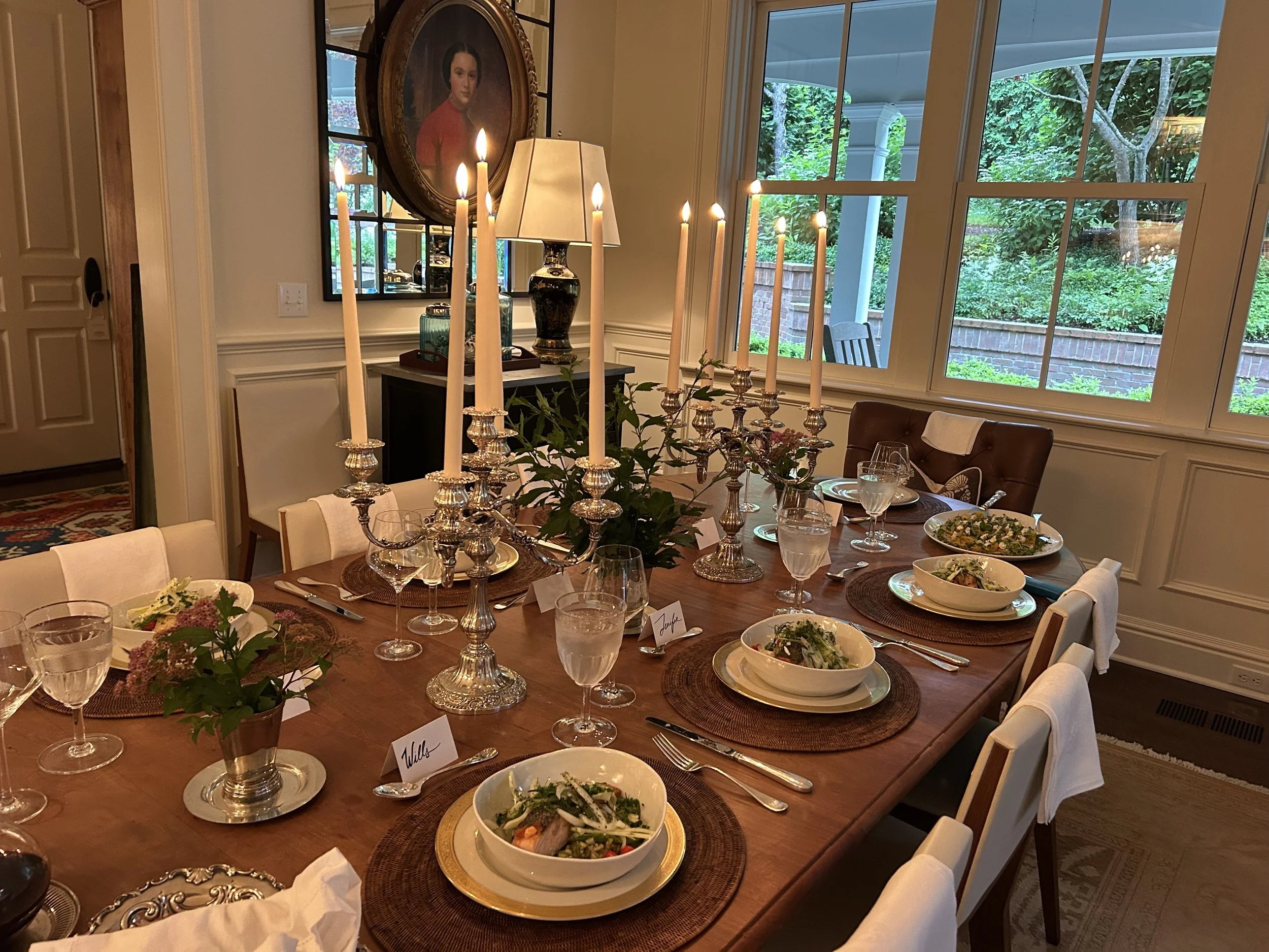 A dining table set for a meal with plates of salad, glasses of water, and silverware. The table has tall candlesticks with lit candles and small flower arrangements. There are chairs around the table, a window with a view of a backyard, and a portrai