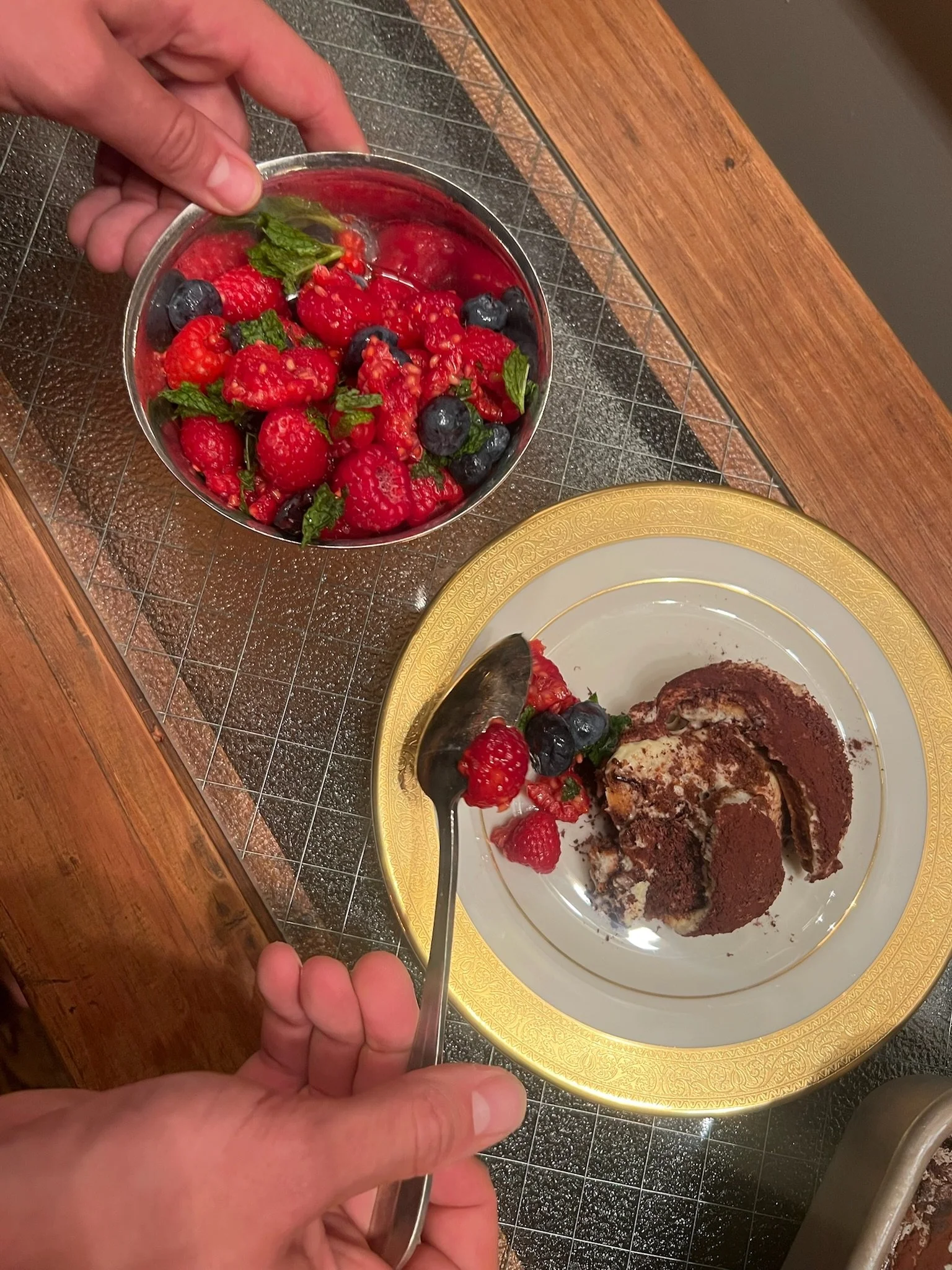 A person is serving fresh mixed berries, including raspberries, blueberries, and mint leaves, onto a plate of tiramisu with a gold Rim.