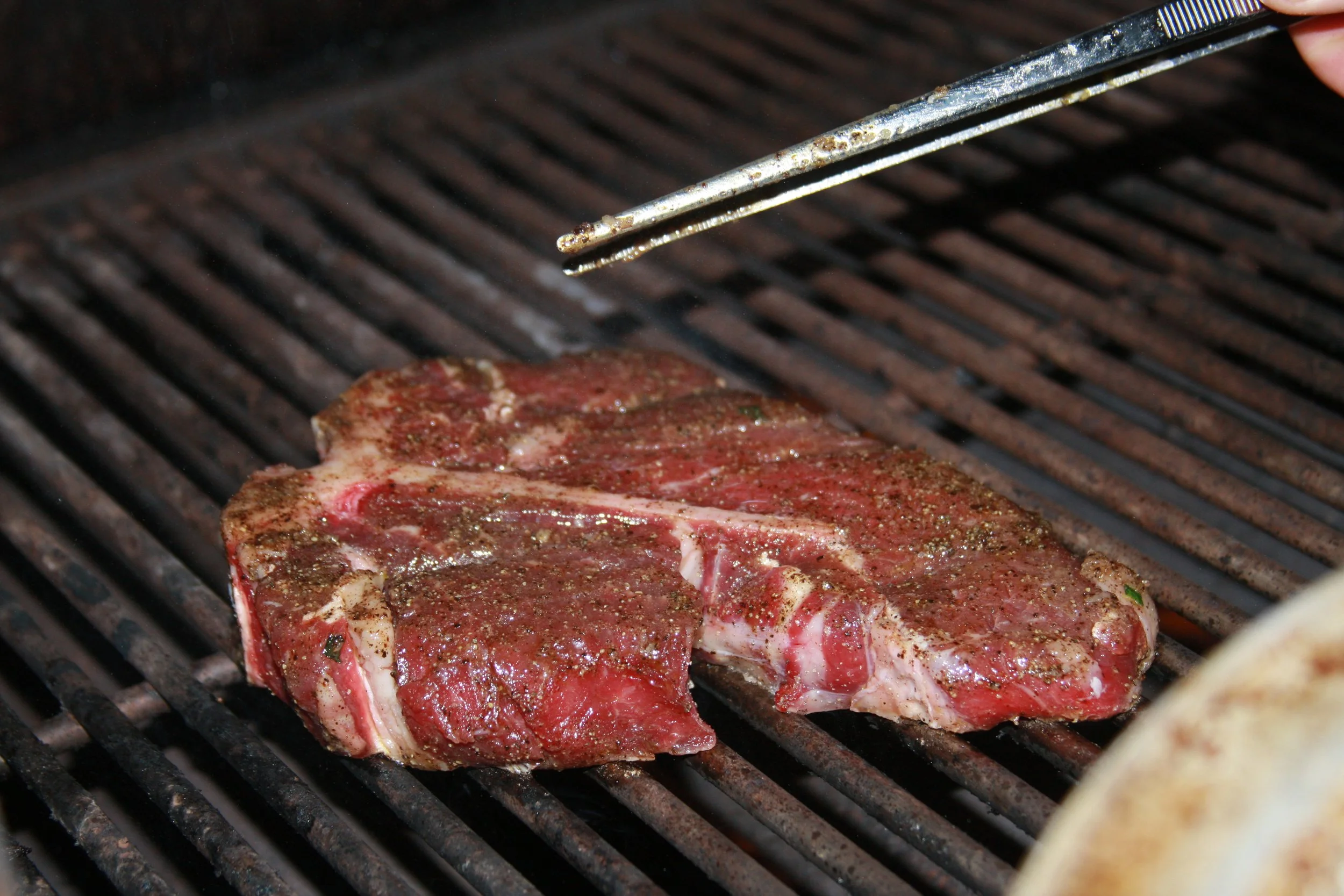 Two raw steaks seasoned with pepper and spices on a grill, with tongs above.