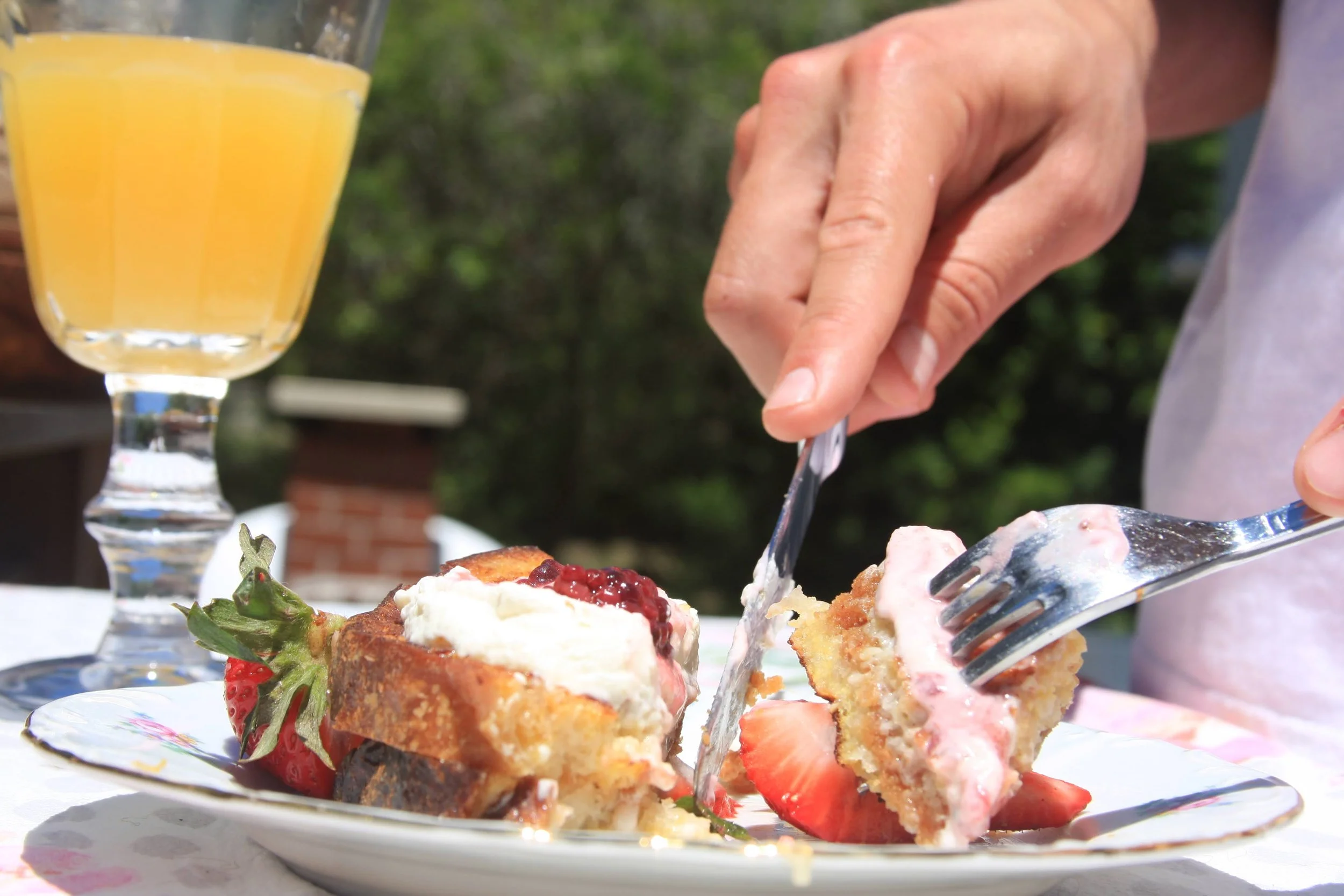 Person cutting a slice of strawberry shortcake with whipped cream and strawberries on top, on a decorative plate. A glass of orange juice in the background.
