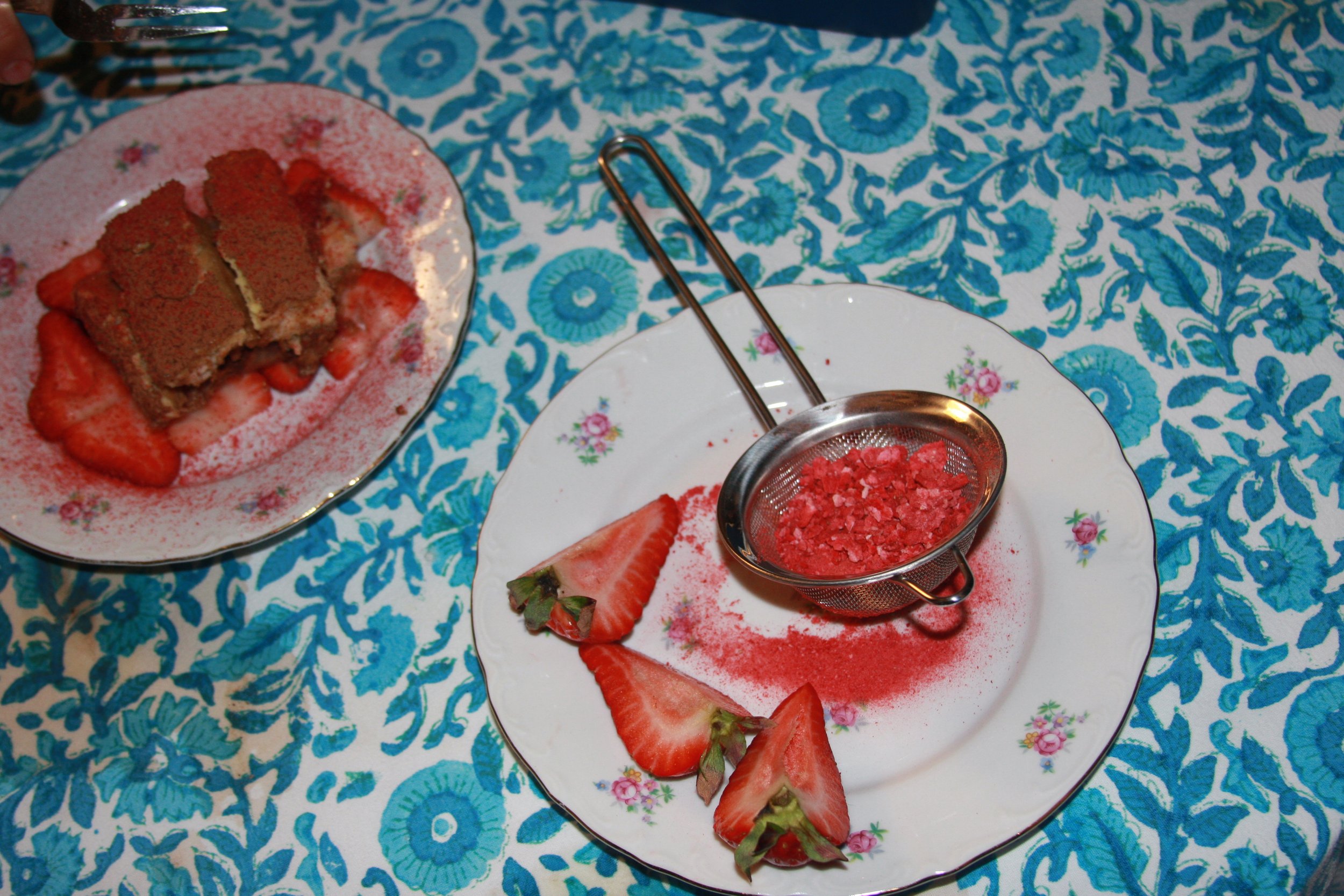 A white floral plate with sliced strawberries, red powder, and a small metal sieve containing crushed red topping. A separate small pink dish holds a layered dessert with strawberries, and the tablecloth has a blue floral pattern.