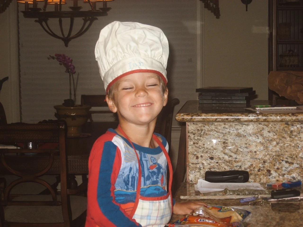 A young boy smiling with eyes closed wearing a chef's hat and apron in a kitchen.