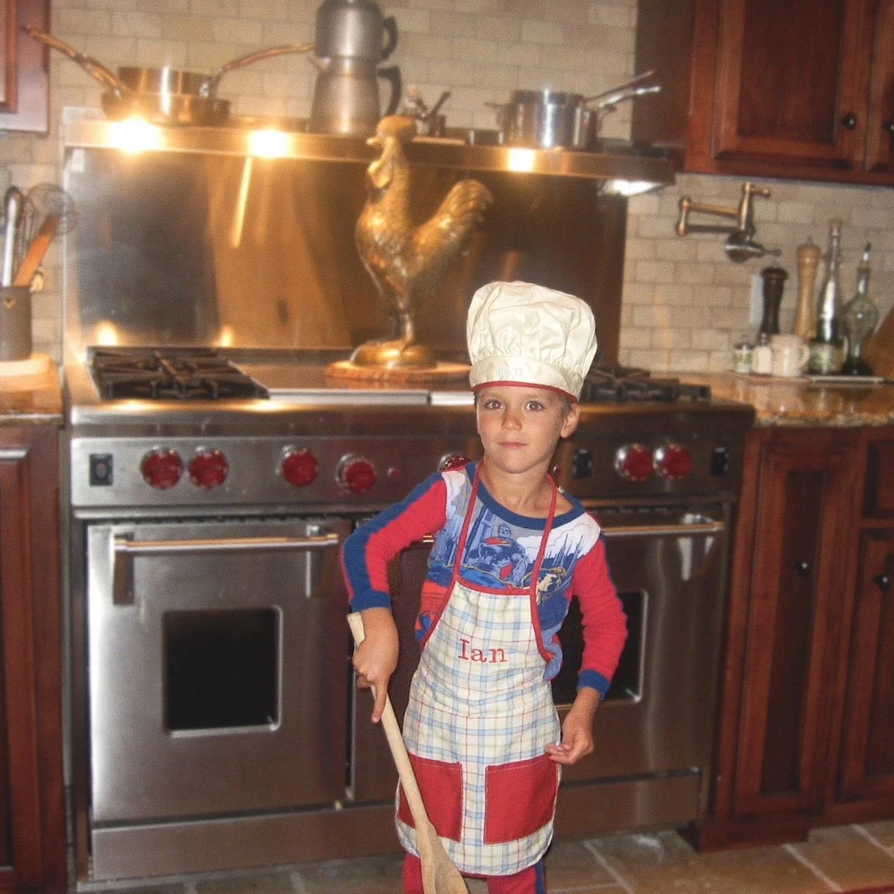 A young boy dressed as a chef with a chef hat and apron, holding a wooden spoon, standing in a kitchen in front of a stove with pots and a decorative rooster.
