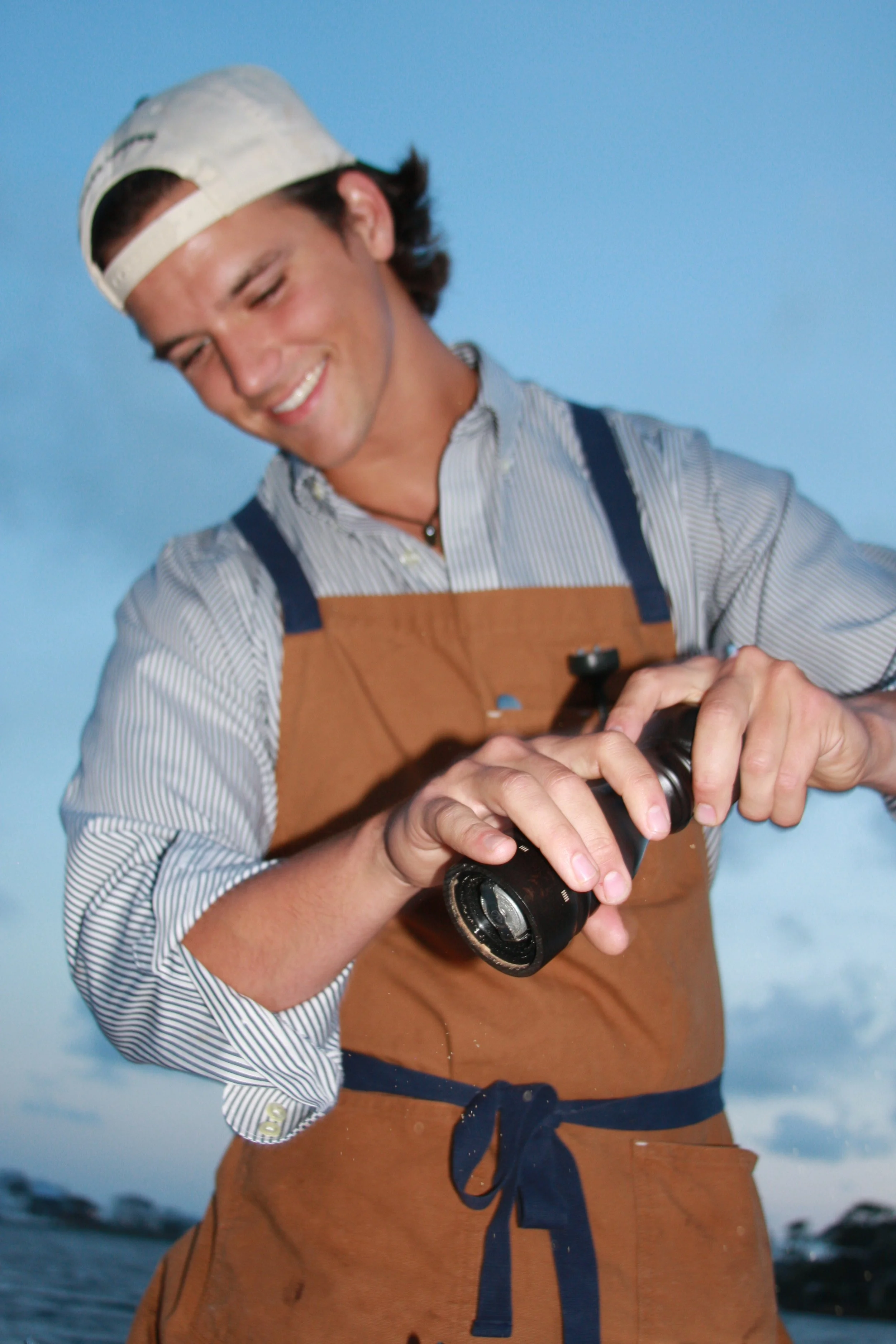 A young man is smiling while holding a camera outdoors during daytime, wearing a striped shirt, a brown apron, and a white cap. The background features a blue sky and water.