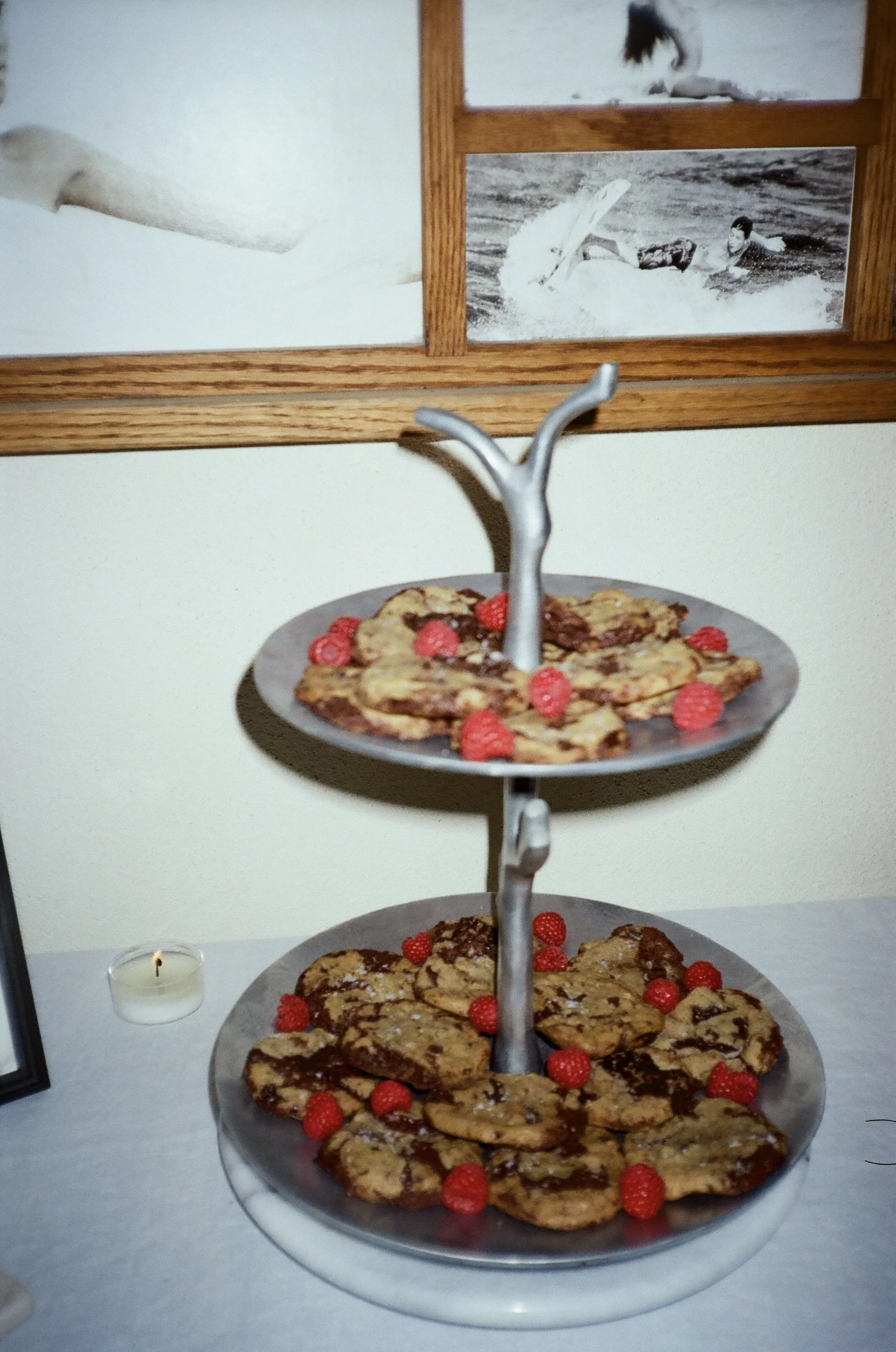 Three-tiered serving tray with chocolate chip cookies and raspberries on each tier, set on a table with a white tablecloth.