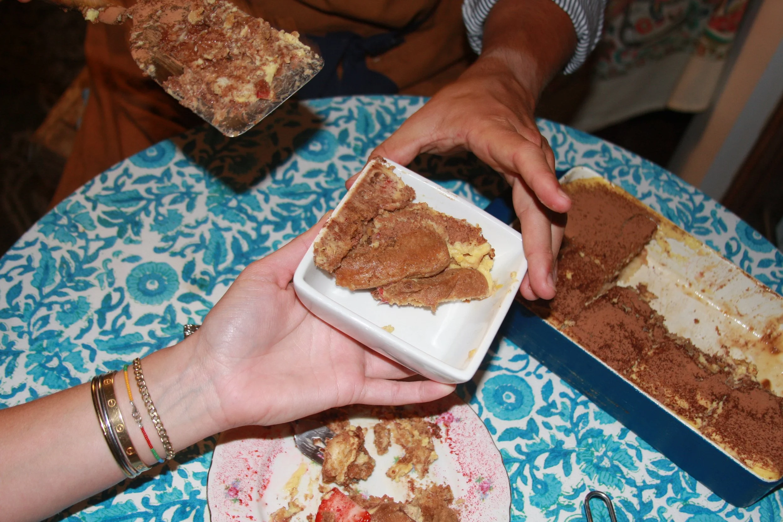 Person holding a small plate with a slice of layered dessert, with a larger tray of the same dessert on the table. The table has a blue and white patterned tablecloth, and other dessert remnants are on a plate. Another person is reaching for the tray