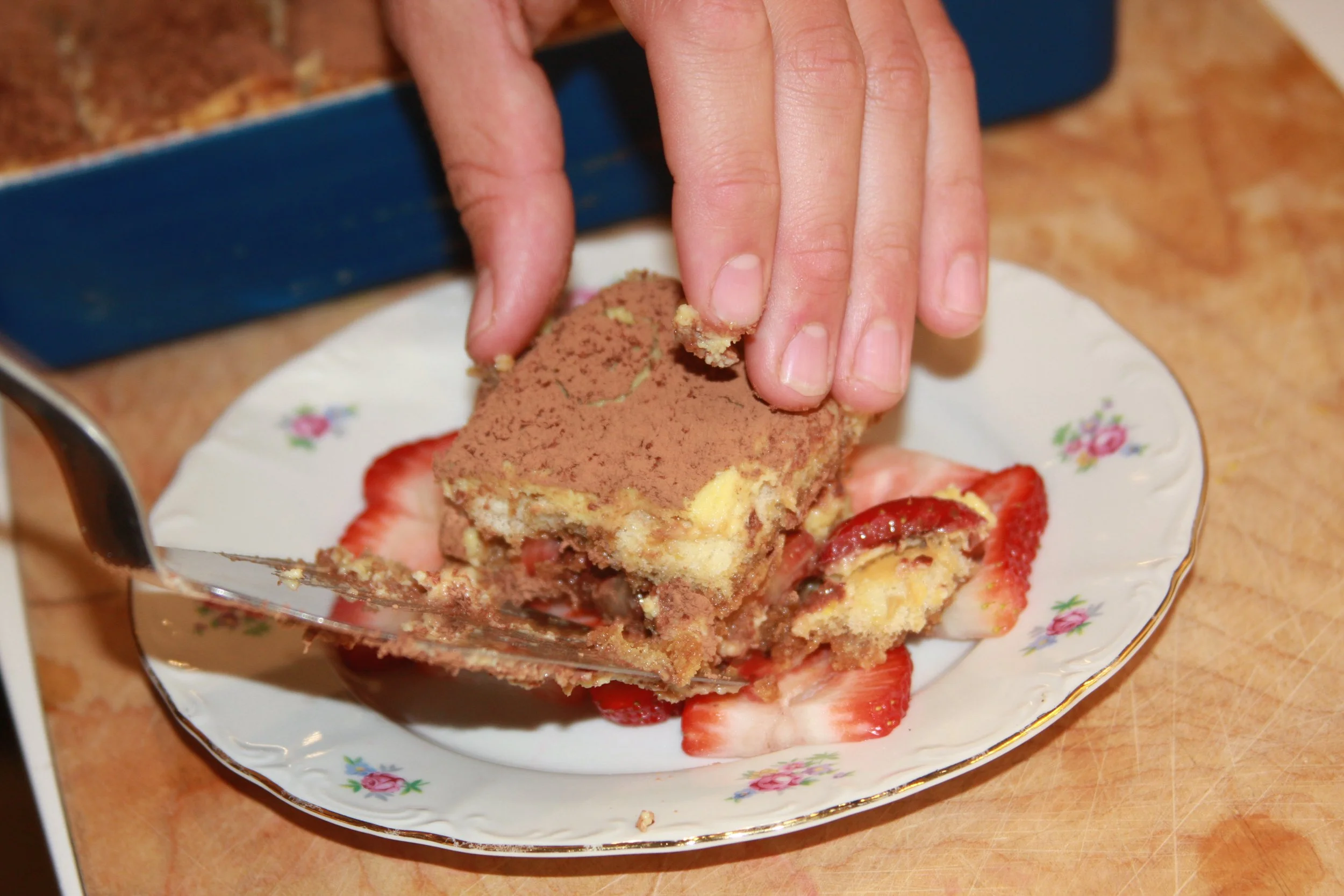 A hand serving a piece of layered dessert with strawberries on a floral plate.