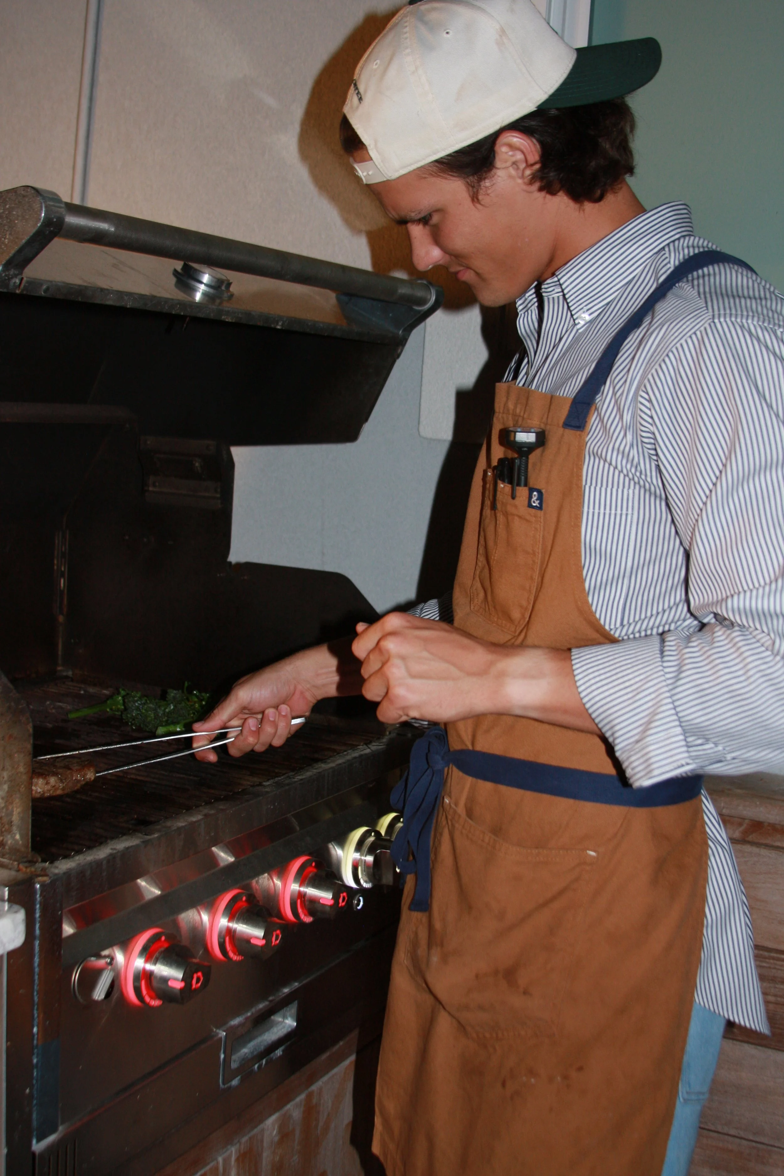 Young man grilling meat and vegetables on a barbecue grill, wearing an apron and a baseball cap.
