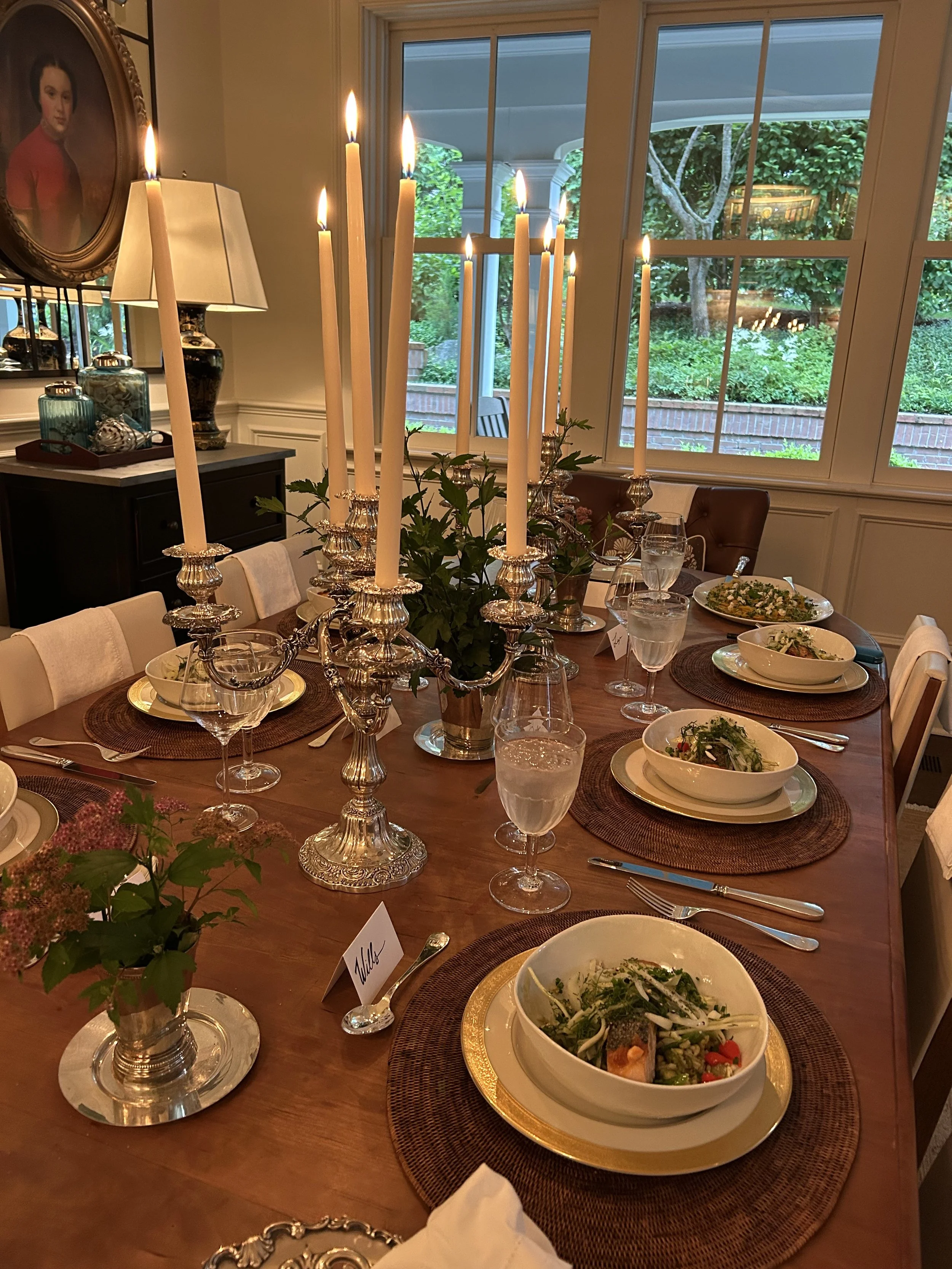 Elegant dining table set with bowls of salad, glasses of water, cutlery, and a large silver candelabrum with lit candles. The table is decorated with small flower arrangements and a name card labeled 'Milds'. Large windows in the background reveal a 