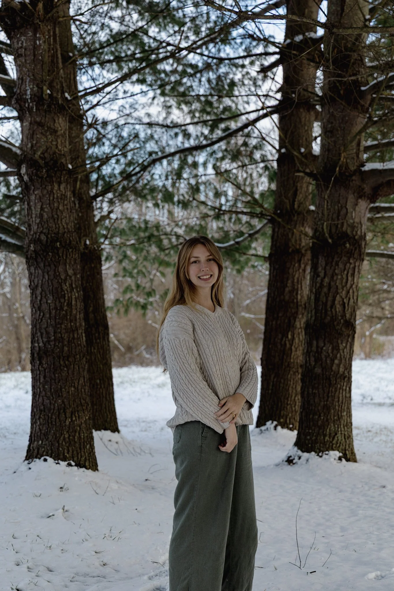A young woman with long blonde hair, wearing a beige sweater and green pants, standing outdoors in a snowy landscape with tall pine trees and a partially cloudy sky.