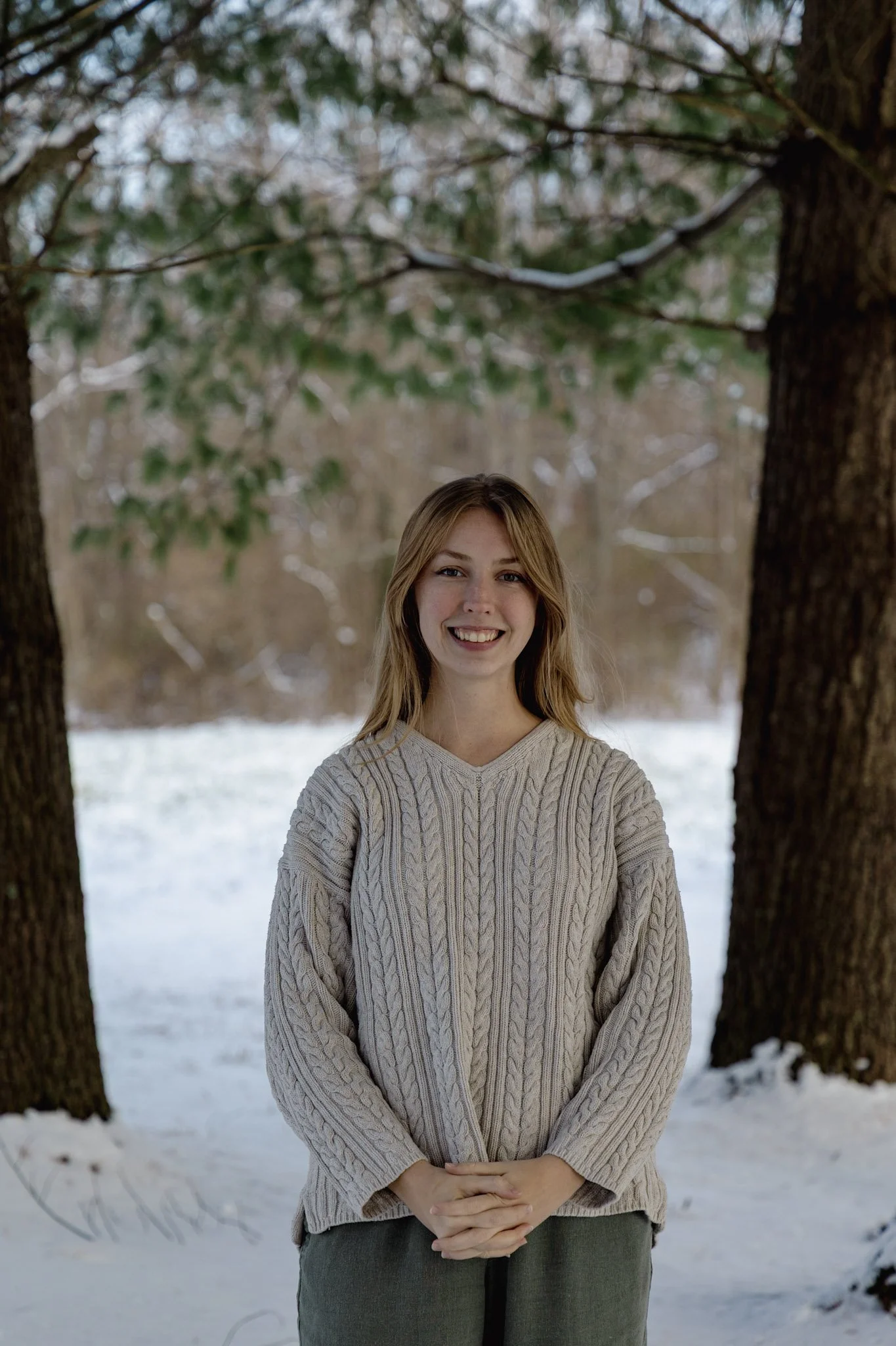 A young woman standing outdoors in a snowy landscape, smiling, with two large trees on either side, wearing a beige cable-knit sweater.
