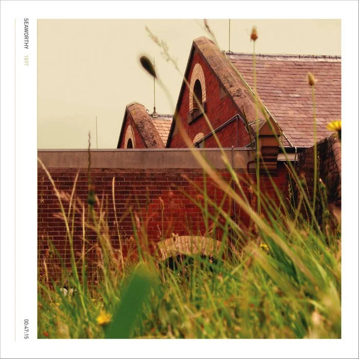 Photo of old brick buildings with triangular roofs, viewed from ground level with grasses and wildflowers in the foreground.