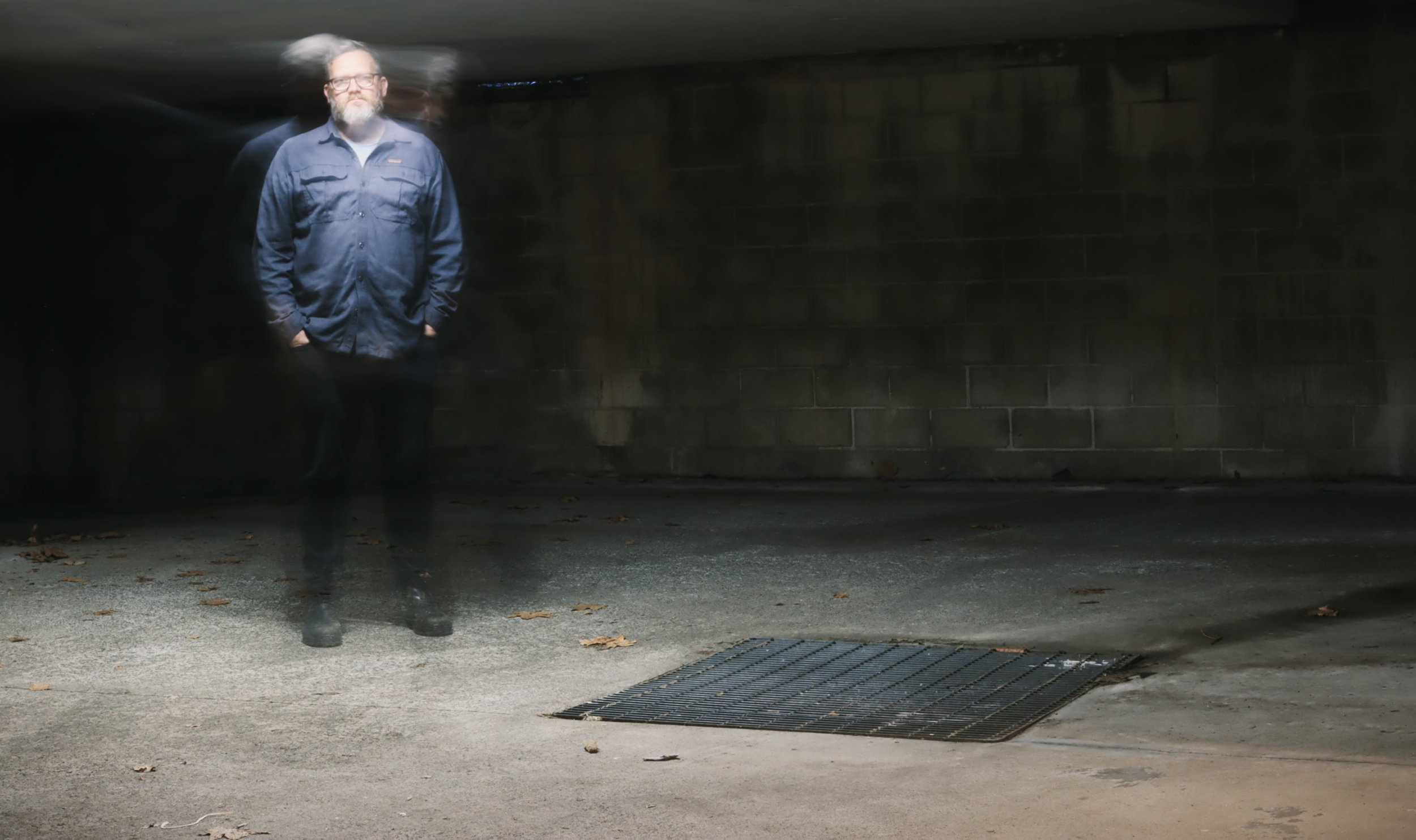 A man standing in an empty parking garage, with motion blur creating multiple images of him and a concrete wall in the background.