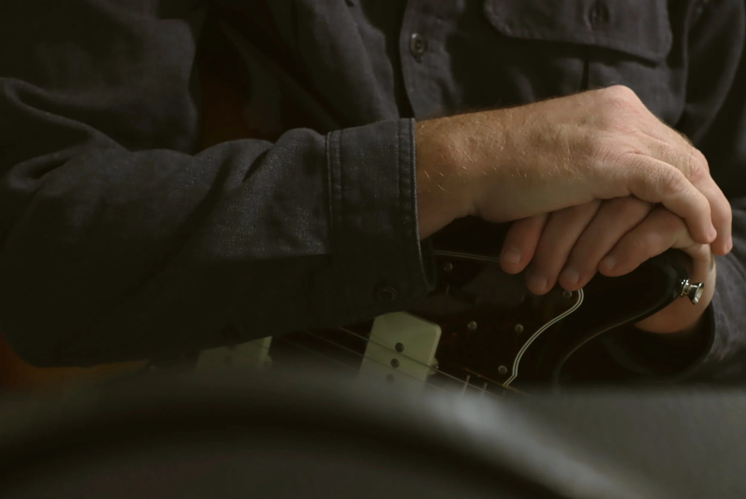Close-up of a person’s hands holding an electric guitar, wearing a dark gray shirt, with the guitar's body partially visible.