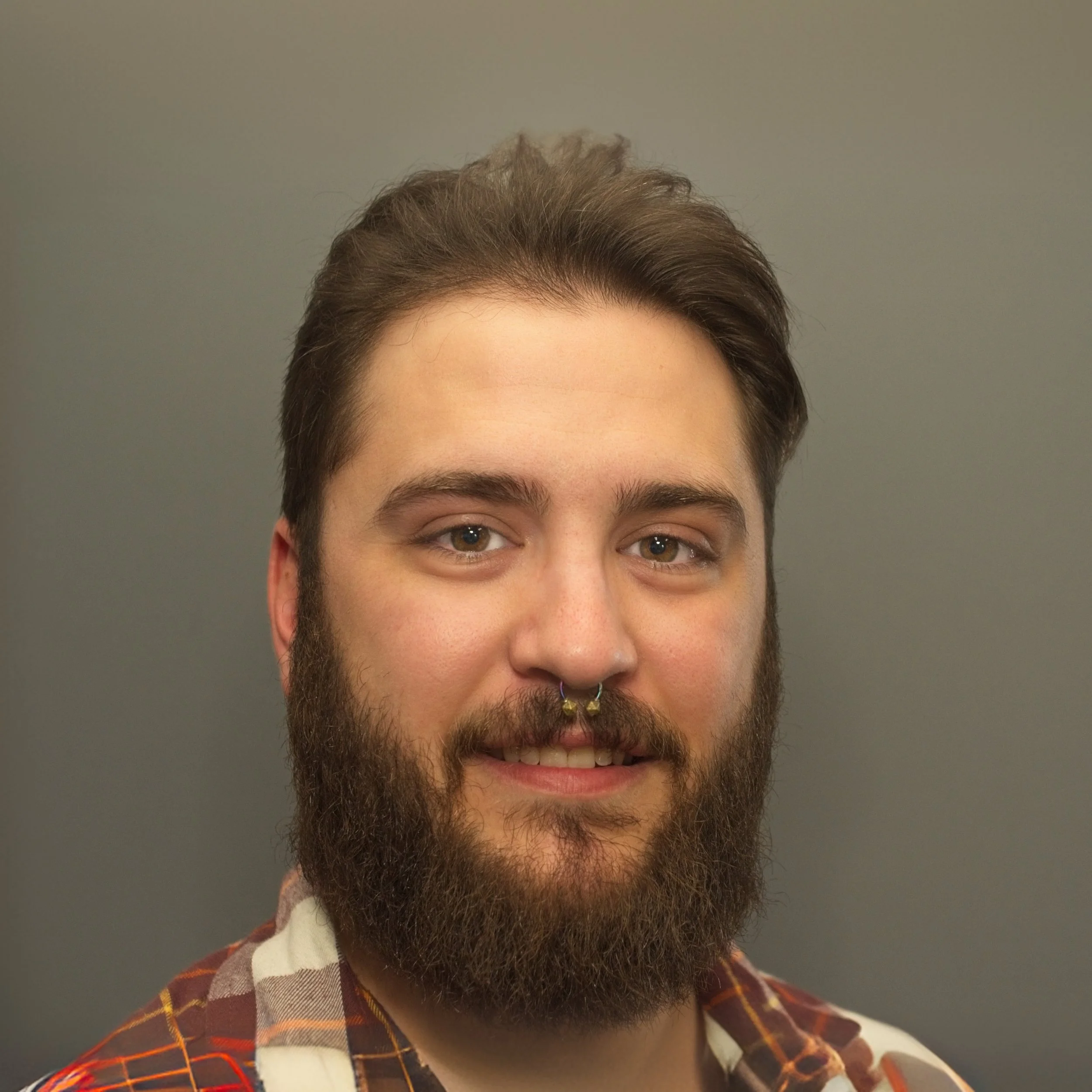 Portrait of a smiling man with brown hair, a beard, and a septum piercing, wearing a patterned shirt against a plain gray background.