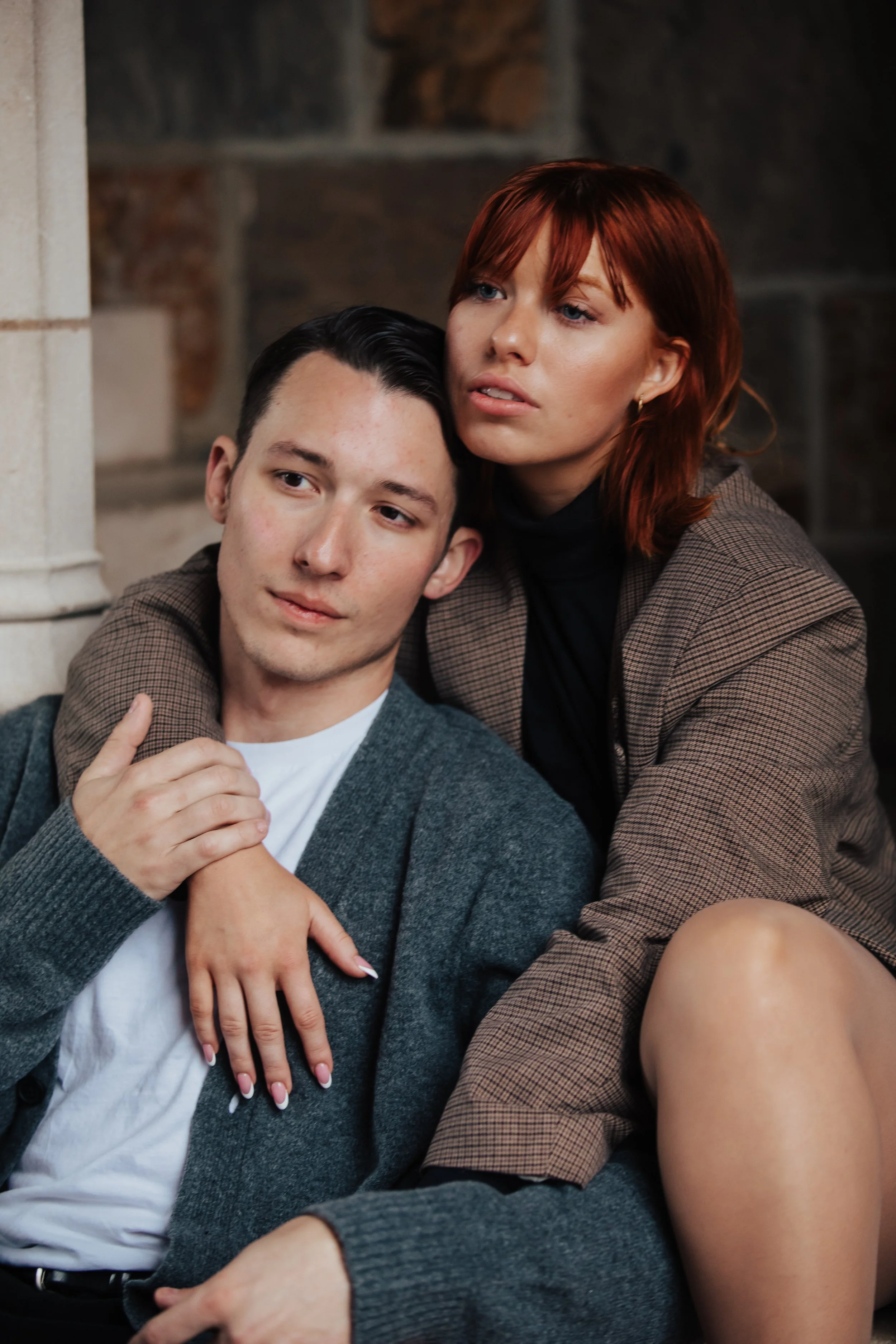 A young man and woman sitting closely together, with the woman resting her head on the man's shoulder and her arm around his neck.