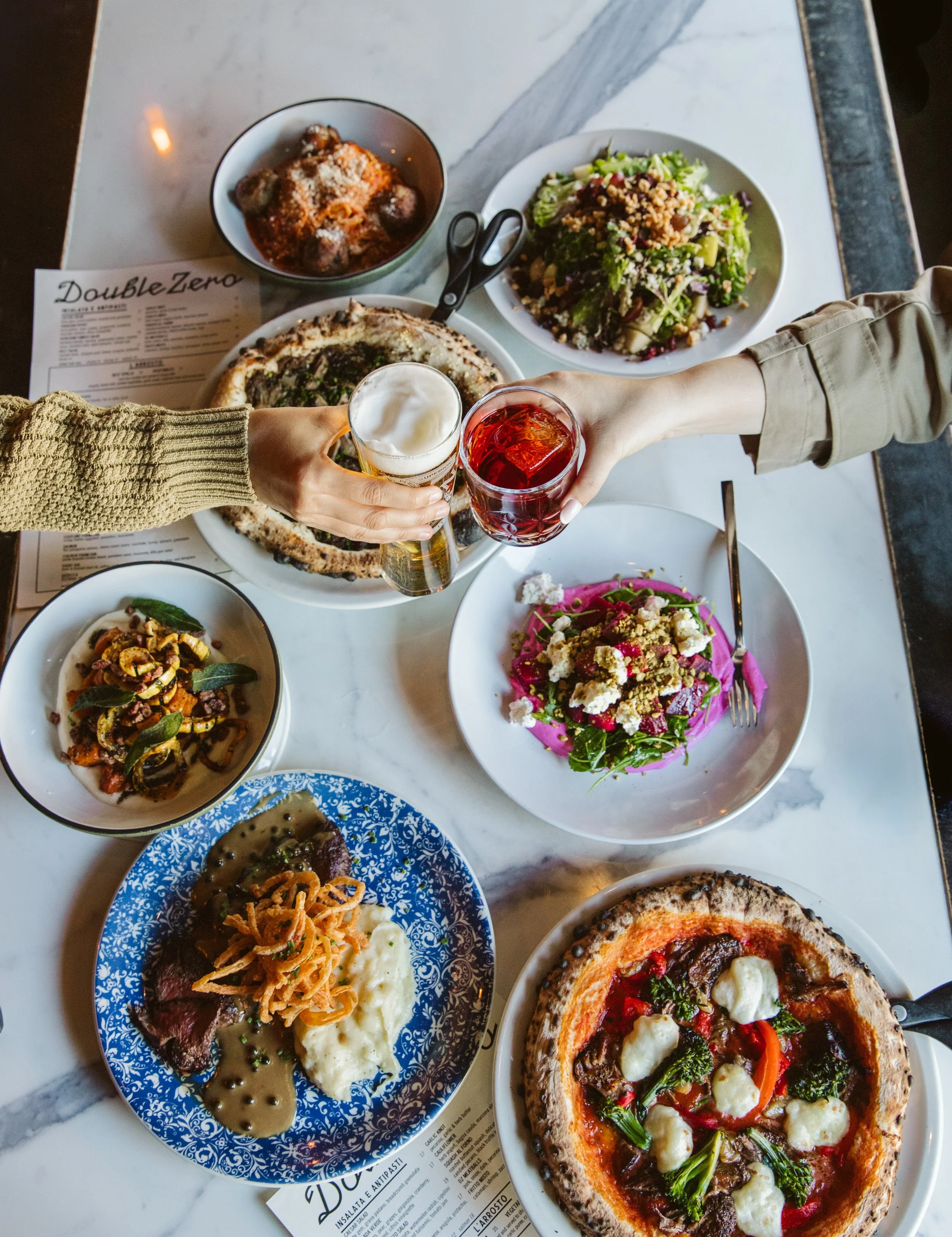 Two people clinking glasses over a table filled with various dishes including pizza, salads, and pasta.