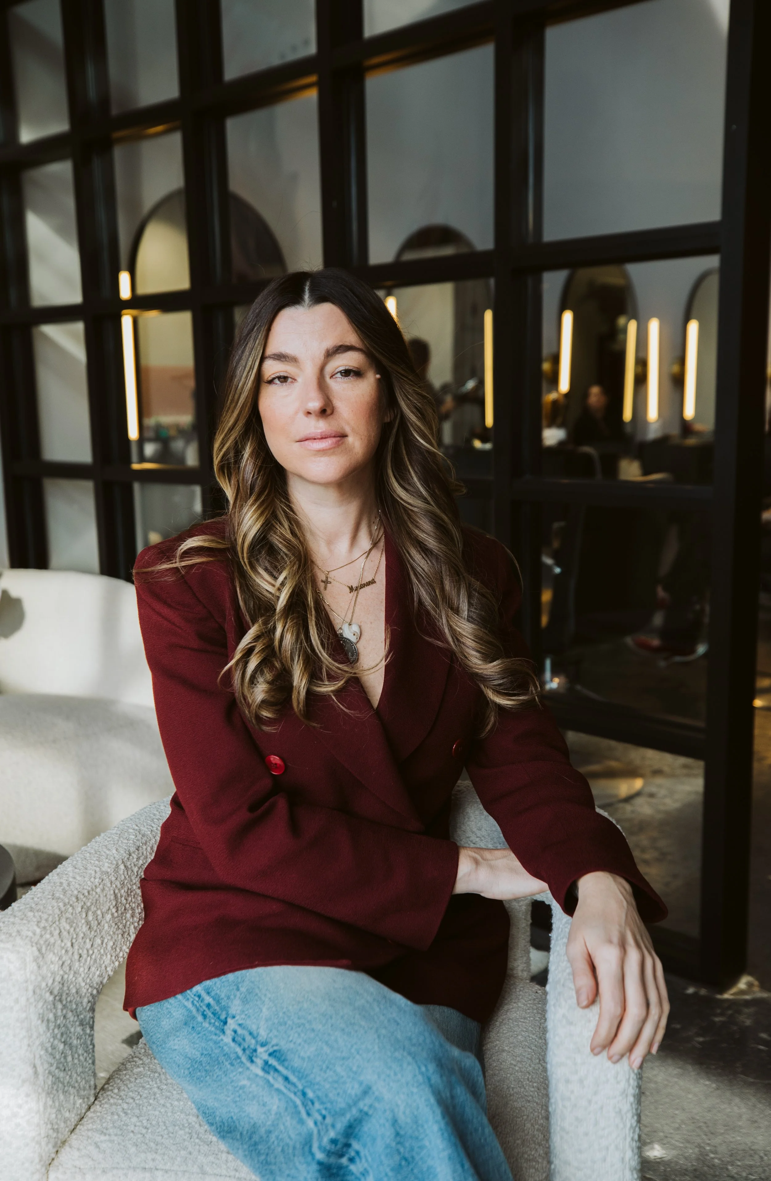 A woman with wavy brown hair wearing a burgundy blazer and blue jeans, sitting on a white textured chair in a modern, upscale interior with black framed glass walls and warm lighting.