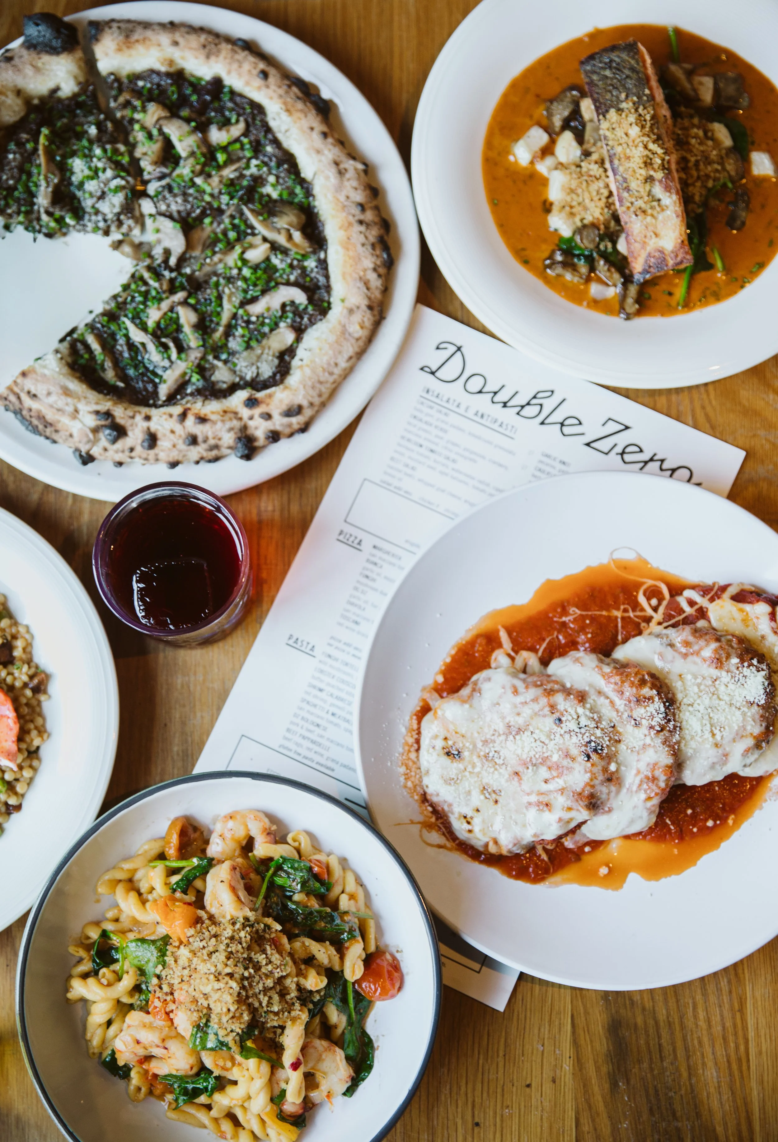 Assorted Italian dishes on a wooden table, including pizza with mushrooms, a piece of fish with sauce, a plate of pasta with shrimp and vegetables, a bowl of seafood pasta, and a plate of baked chicken Parmesan with tomato sauce and cheese, with a gl