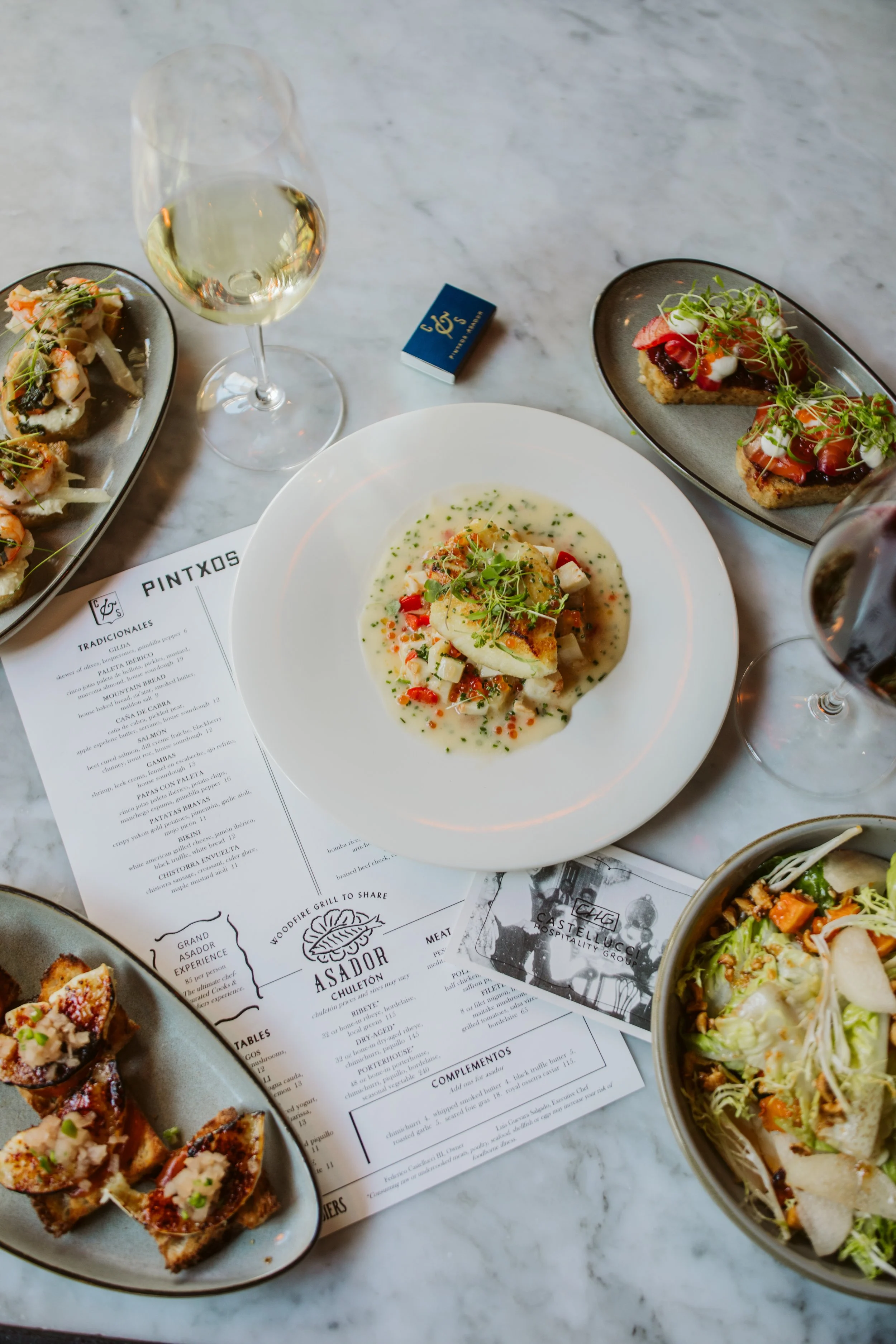 A table with plates of food, glasses of white wine, and menus, showcasing a variety of appetizers and main courses at a restaurant.