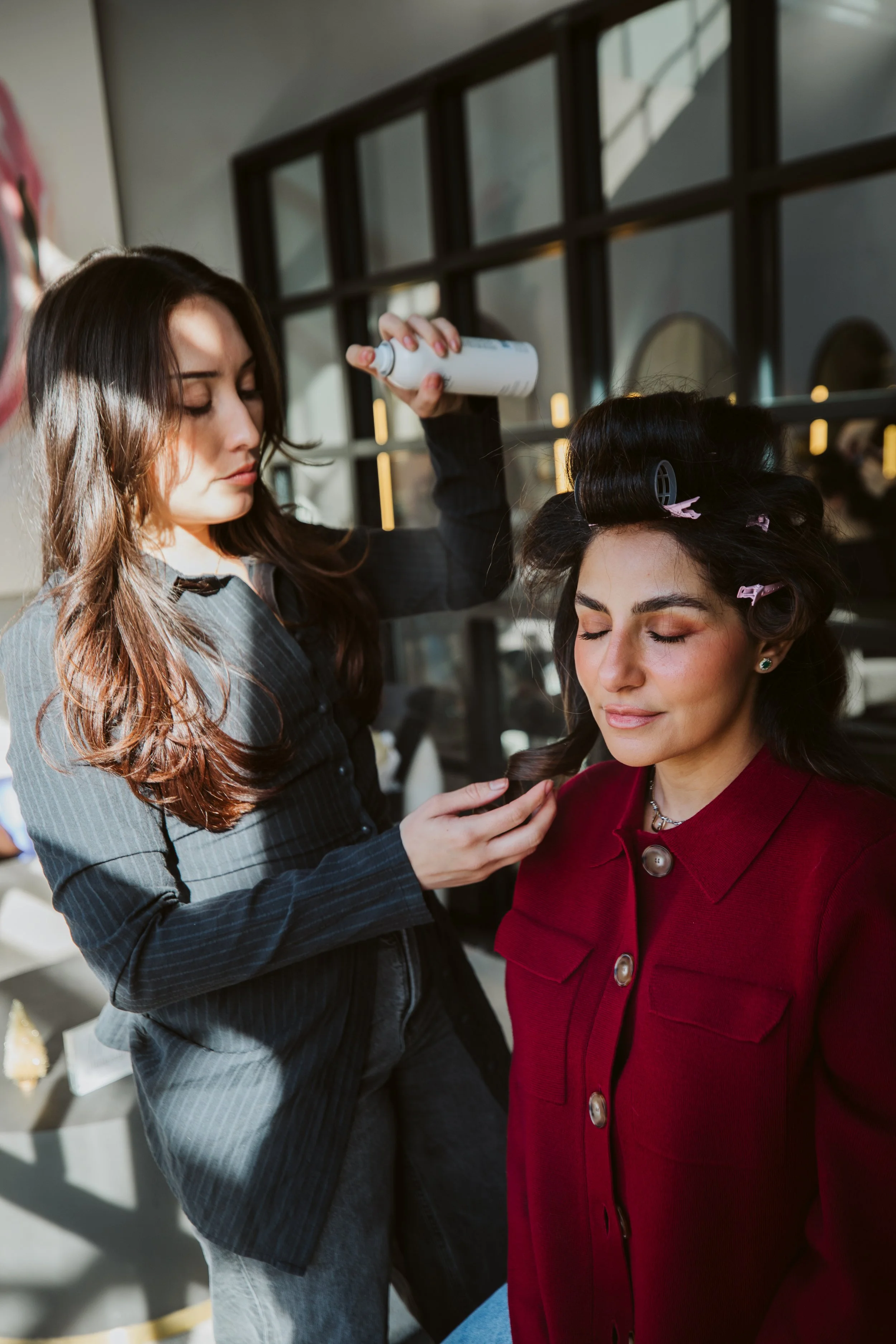 A hairdresser styling a woman's hair with rollers and spray in a salon.