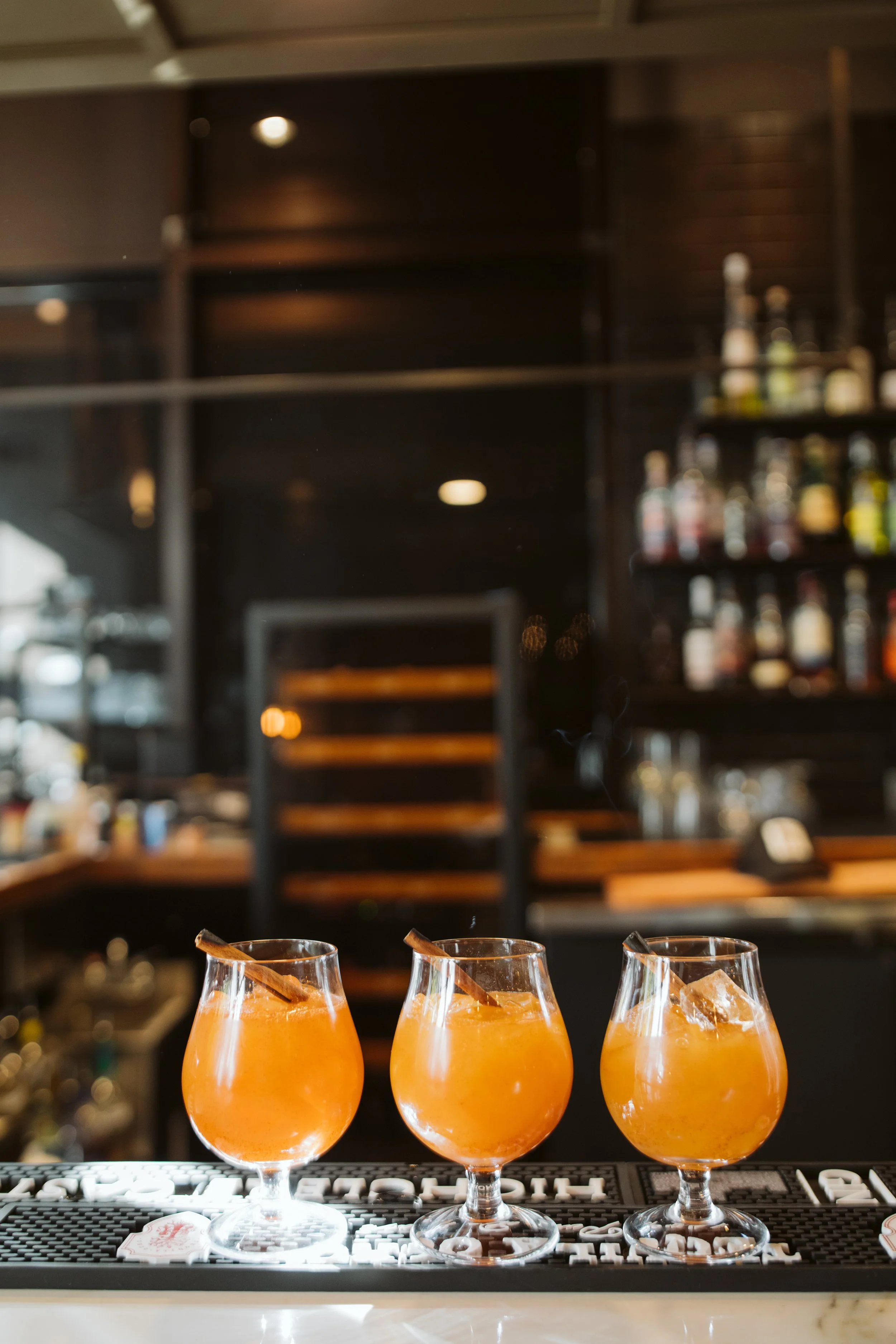 Three orange-colored cocktails with cinnamon sticks and ice cubes on a bar mat at a bar.