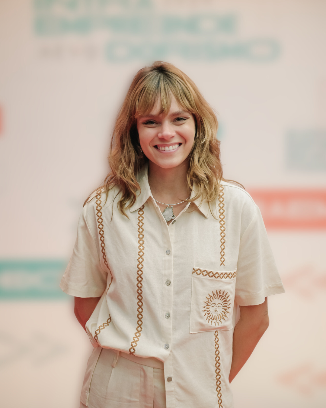 Jovem sorridente com cabelo castanho ondulado, usando uma blusa clara com detalhes de linha e um colar com pingente de mapa.