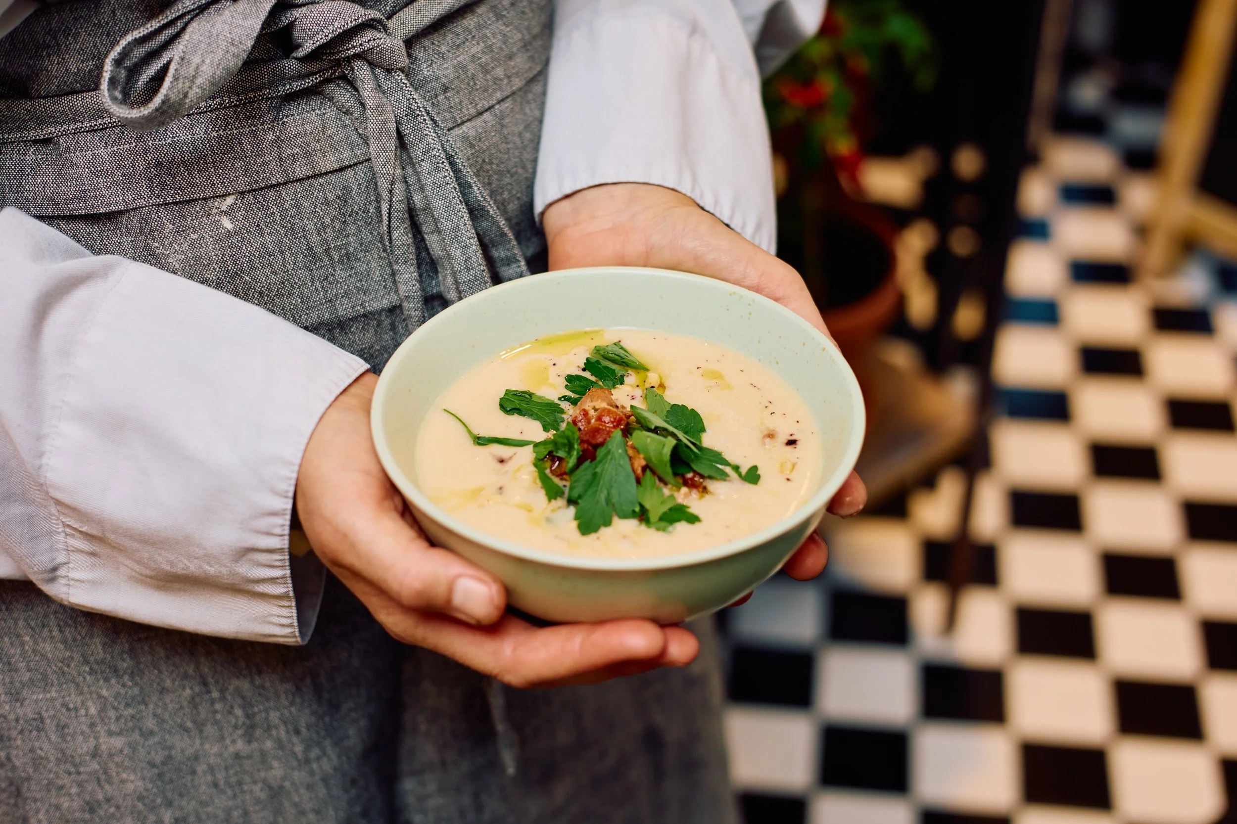 En person som holder en skål med blomkålsuppe, toppet med bacon, pinjekjerner og urter