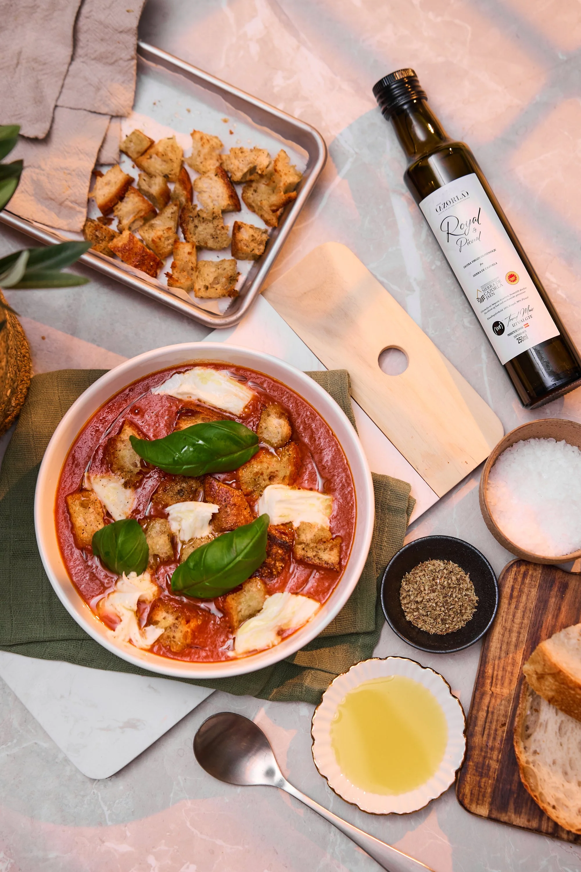 Overbilde av tomatsuppe med mozarella, hjemmelaget krutonger og basilikum på et fjøl på et bord med olje, salt og servise.