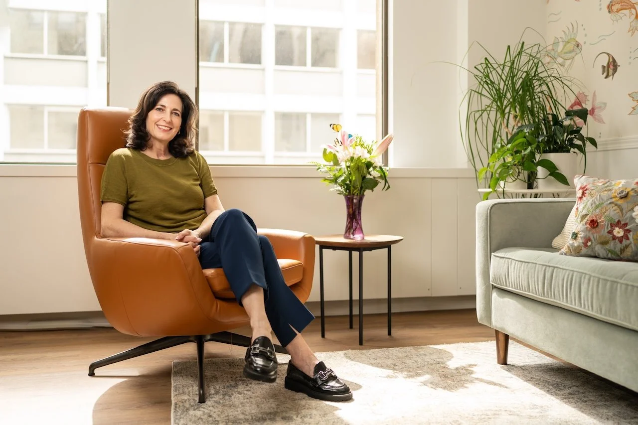 A woman sitting in an orange armchair in a bright room with large windows, a small side table with a flower vase, and a white couch with floral pillows.