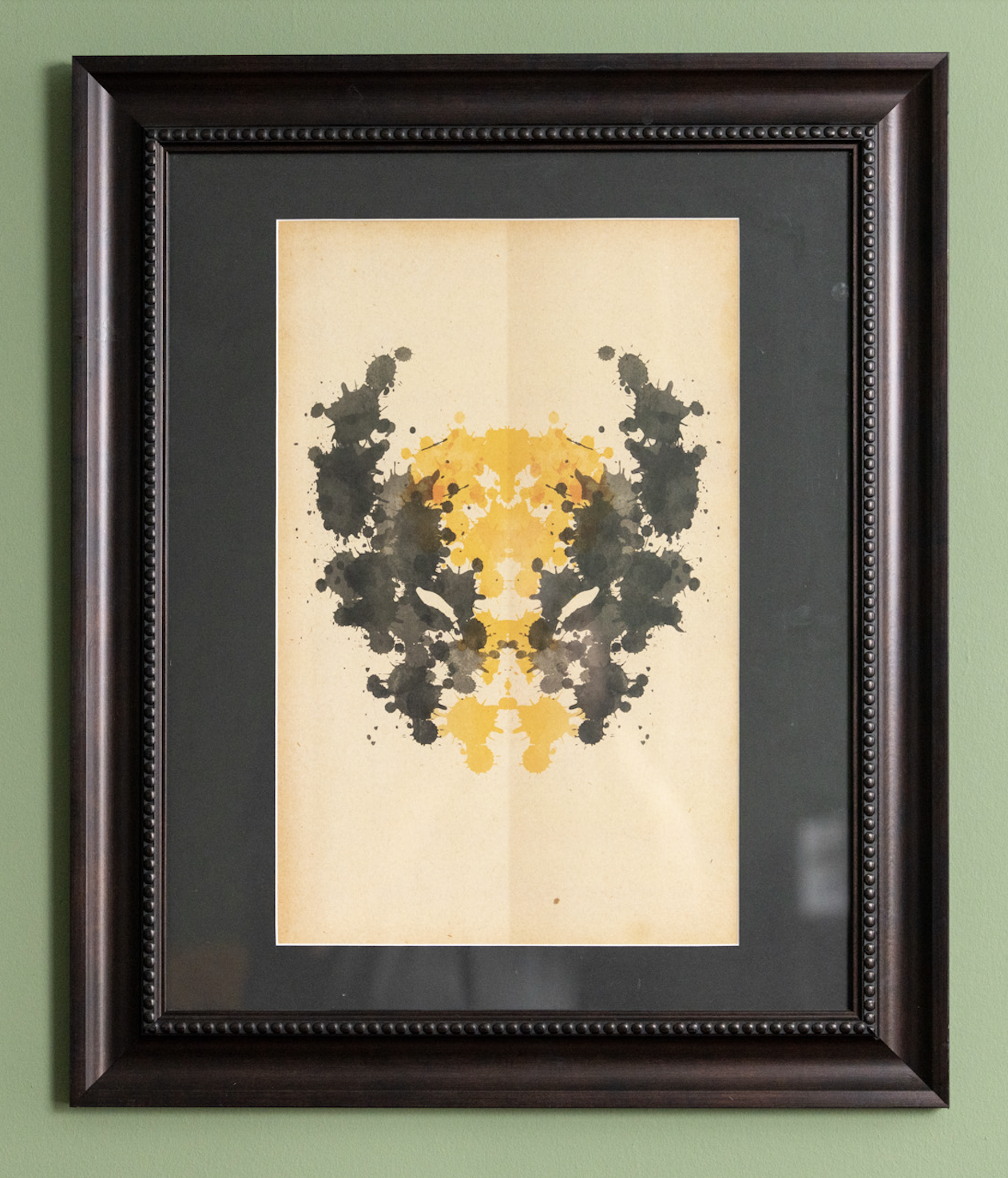 A framed abstract inkblot artwork with black and yellow splashes on beige paper, hanging on a green wall.