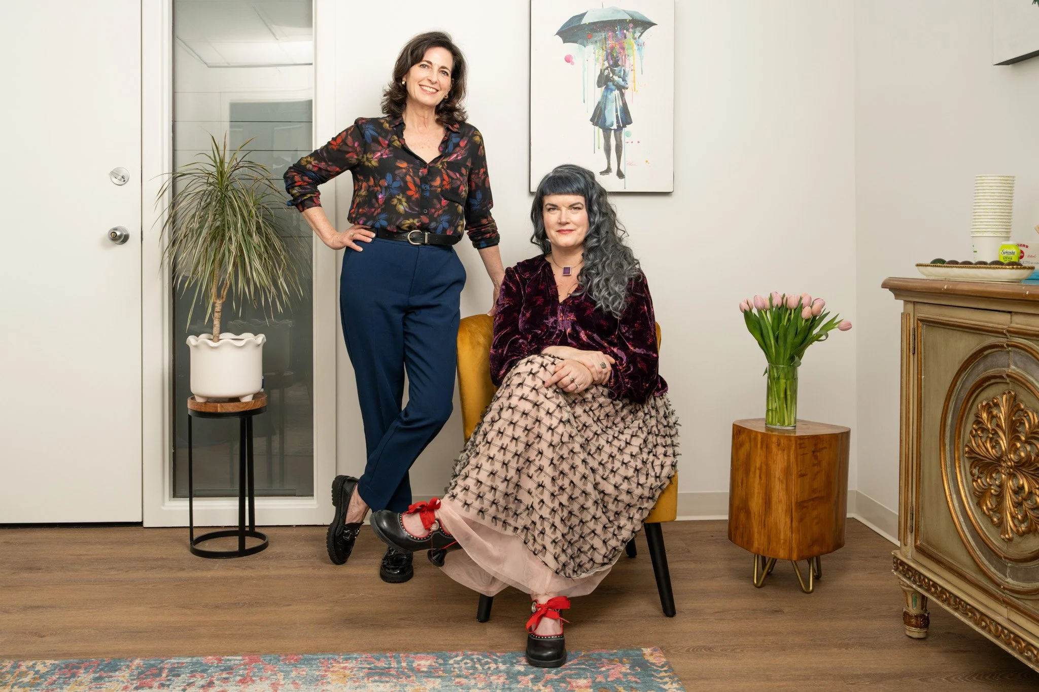 Two women in a room with minimalist decor, one sitting on a yellow chair and the other standing next to her. The woman on the left has shoulder-length dark hair, wears a floral blouse and blue pants. The seated woman has long, curly, gray hair, wears a velvet top and plaid skirt with red shoes. There is a tall potted plant, a vase of pink tulips, a wooden sideboard, and a painting of a girl with an umbrella on the wall.