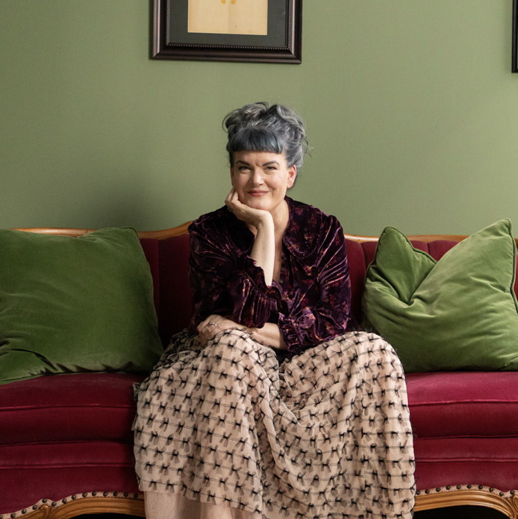 Woman with gray hair sitting on a red velvet couch with green pillows, wearing a velvet top and a patterned skirt, smiling with her hand resting on her chin, in a room with green walls and framed artwork.