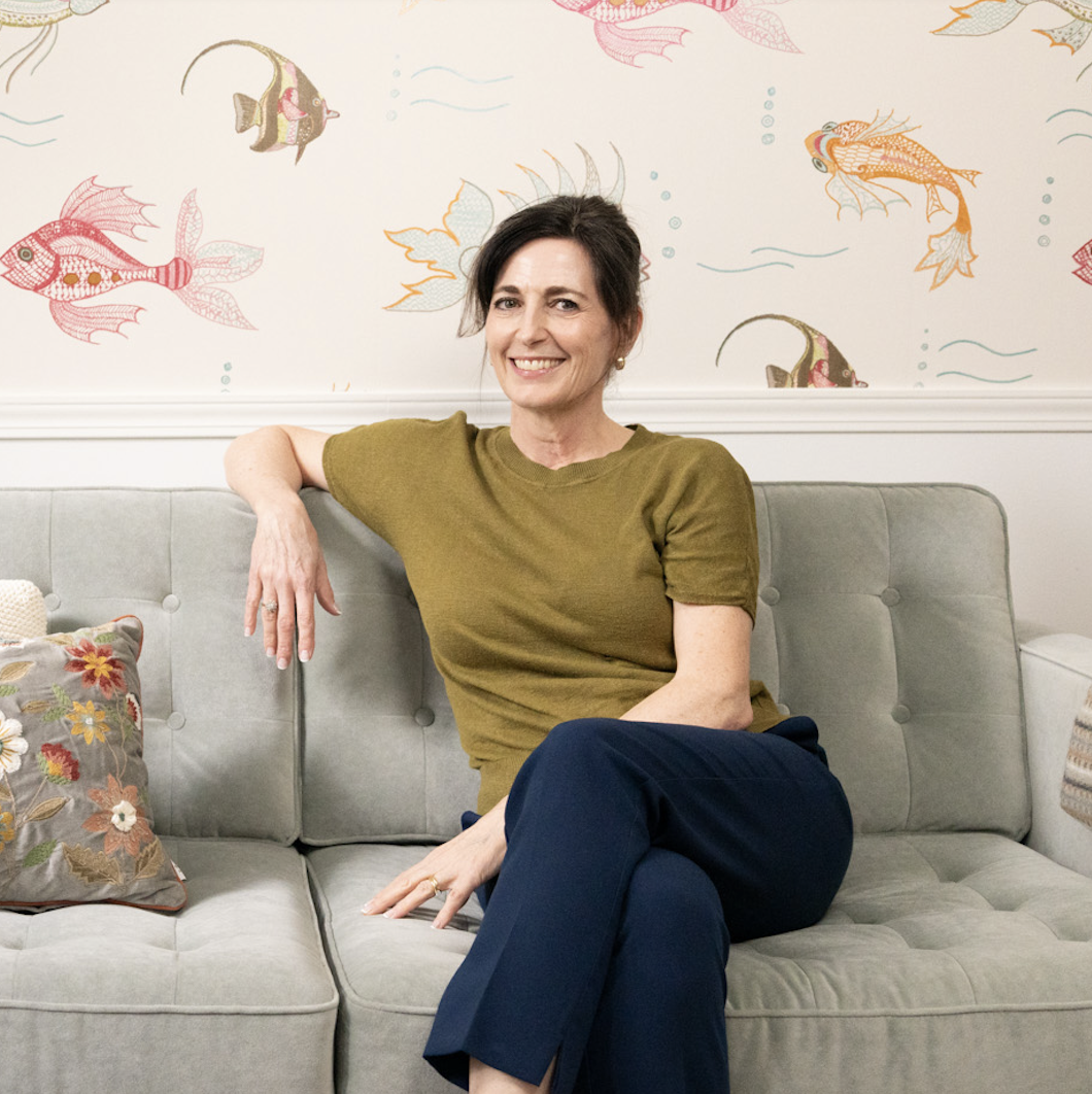 A woman with short dark hair, wearing an olive green T-shirt and navy pants, sitting on a light gray couch in front of a wall with colorful fish and ocean-themed illustrations, smiling at the camera.