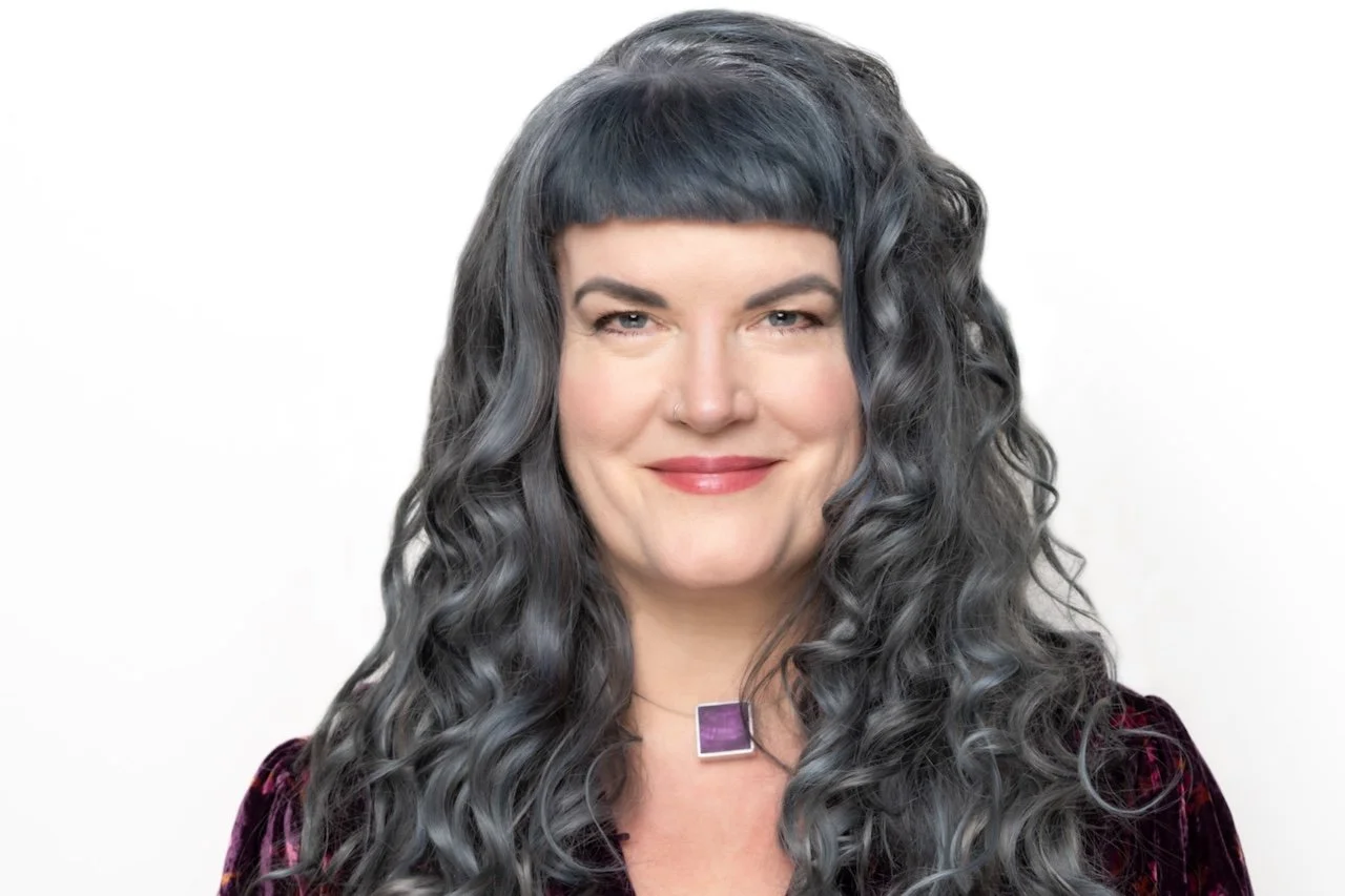 A woman with long, curly, dark gray hair and a blunt fringe, smiling slightly, wearing a dark purple velvet top and a square purple necklace against a plain white background.