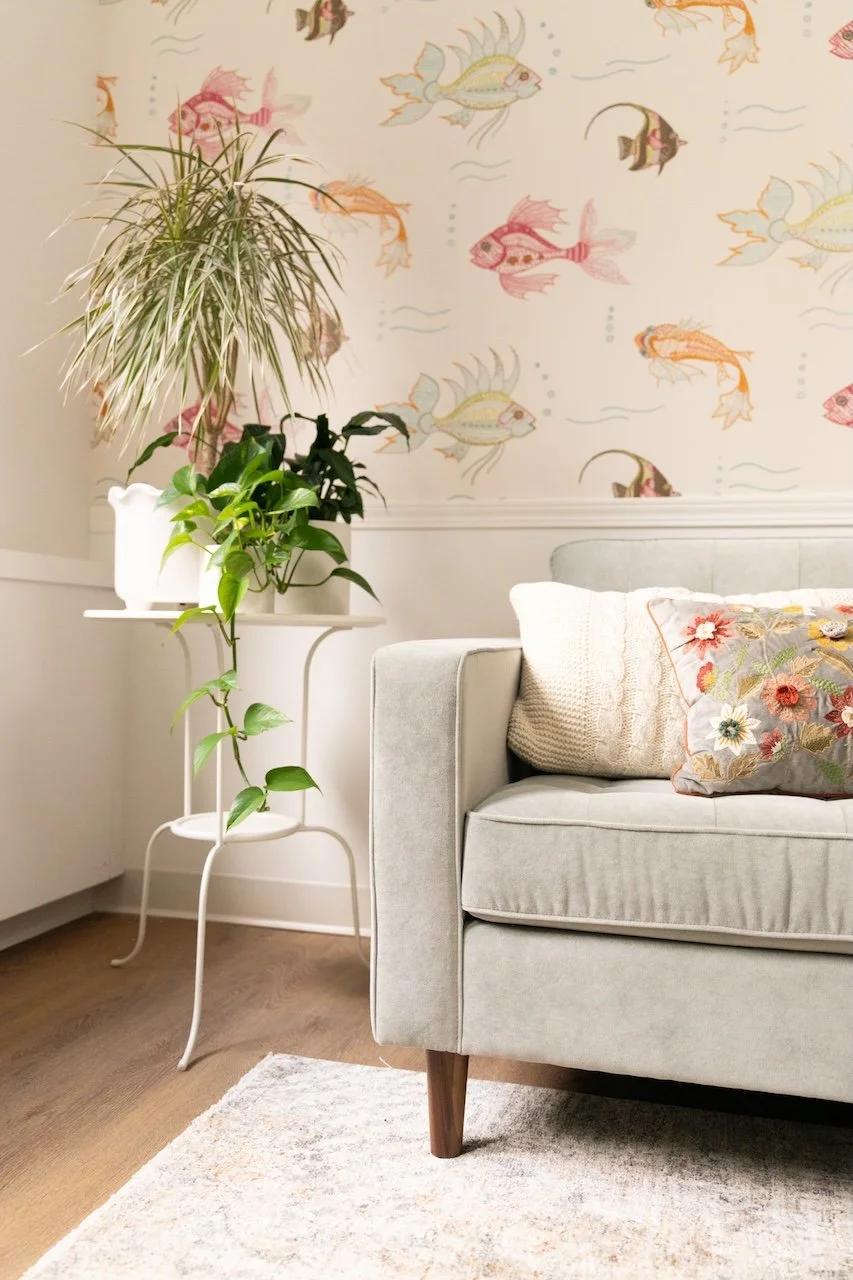 Living room with a light gray sofa, floral throw pillow, cream knitted pillow, white side table with green houseplants, and wallpaper with colorful fish illustrations.