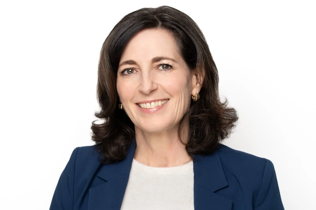 Professional woman with shoulder-length dark hair smiling, wearing a navy blazer and white top, against a white background.