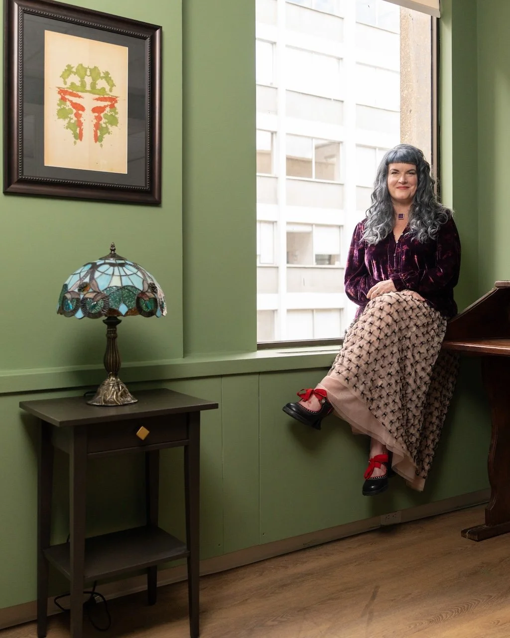 A woman with gray wavy hair sitting on the window ledge of a green-walled room, smiling at the camera. She is wearing a dark velvet top, patterned wide-leg pants, and black shoes with red bows. Next to her is a small table with a Tiffany-style stained glass lamp, and a framed artwork with a green and orange abstract design hangs on the wall.
