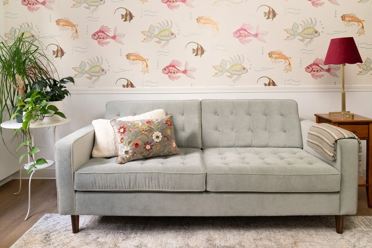 Living room with a light gray tufted sofa, floral pillow, striped blanket, wooden side table with a red lampshade, potted plants on a white stand, and a wallpaper with colorful fish pattern.