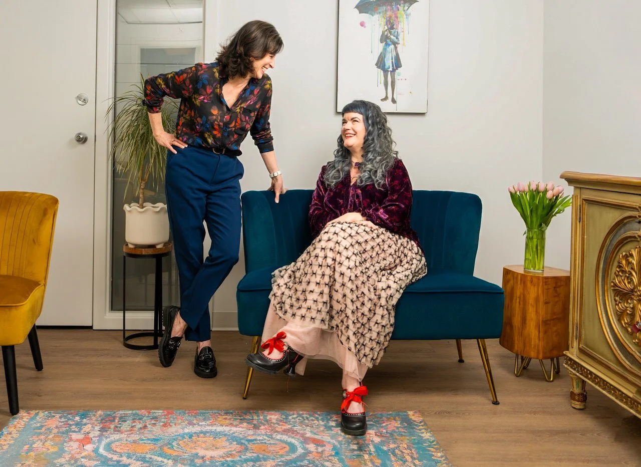 Two women sharing a friendly moment in a living room, one sitting on a blue couch with gray curly hair and the other standing and leaning on the couch with brown hair, both smiling and engaging in conversation.