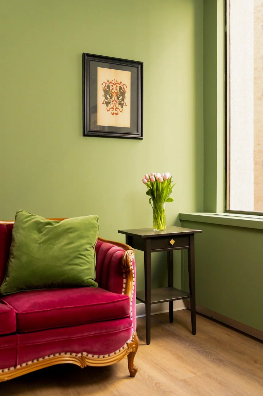 A cozy corner with a pink velvet vintage sofa with a green cushion, a black side table with a glass vase of pink tulips, a green wall with a framed artwork, and a large window letting in natural light.