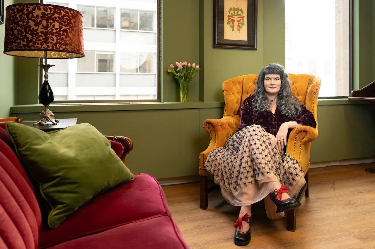 A woman with long gray curly hair sitting in a yellow armchair in a room with green walls and wooden floors. There is a window behind her with a vase of pink tulips on the windowsill and a framed piece of art on the wall. A red sofa with a large green pillow is partially visible in the foreground, and a black table lamp with a patterned shade is on a side table next to the sofa.