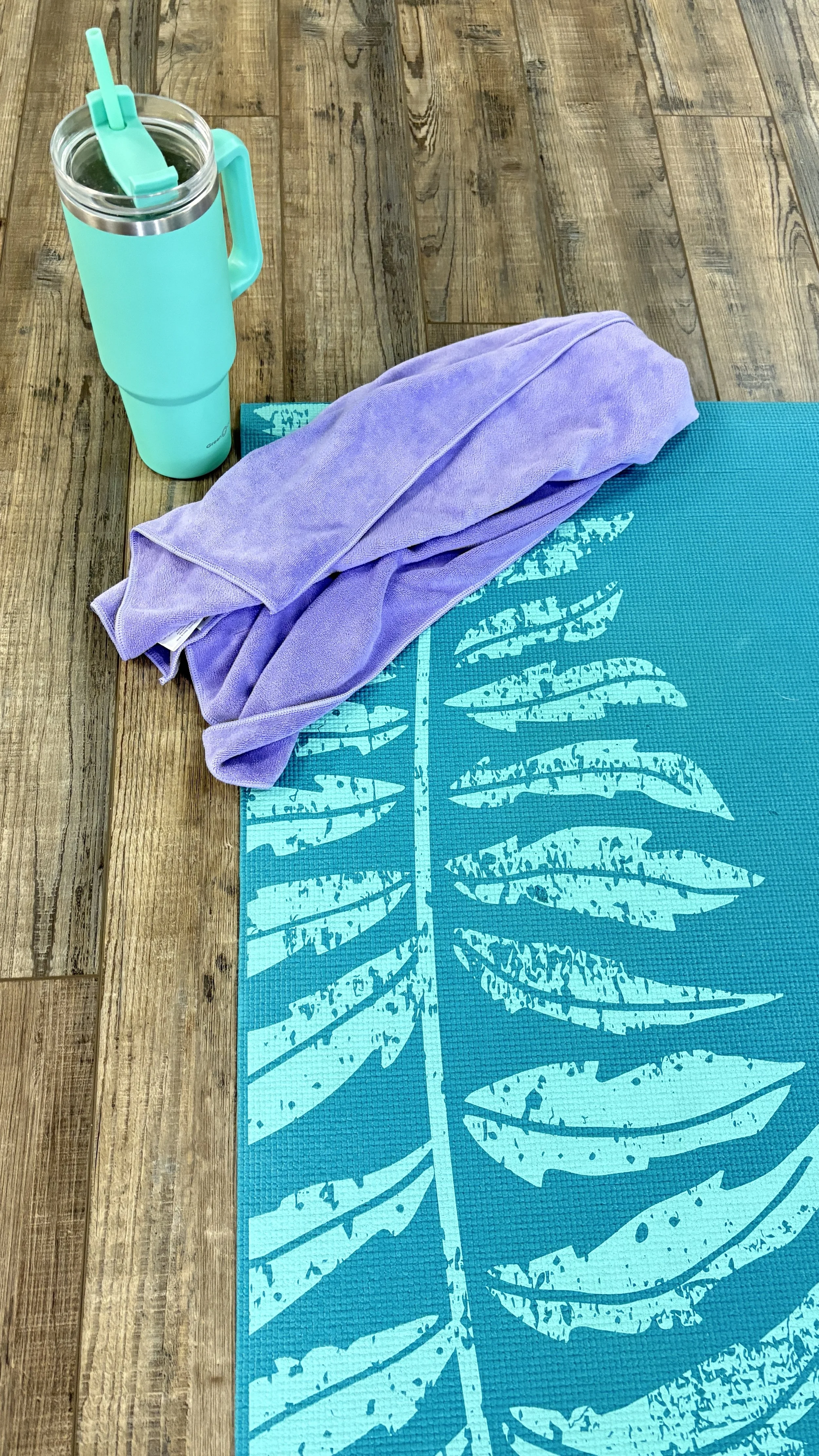 A yoga mat with a leaf pattern on a wooden floor, a folded purple towel, and a teal water bottle with a straw.